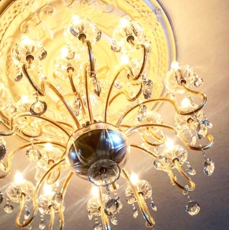 A chandelier with multiple crystal prisms and bright lights hanging from a ceiling with ornate molding.