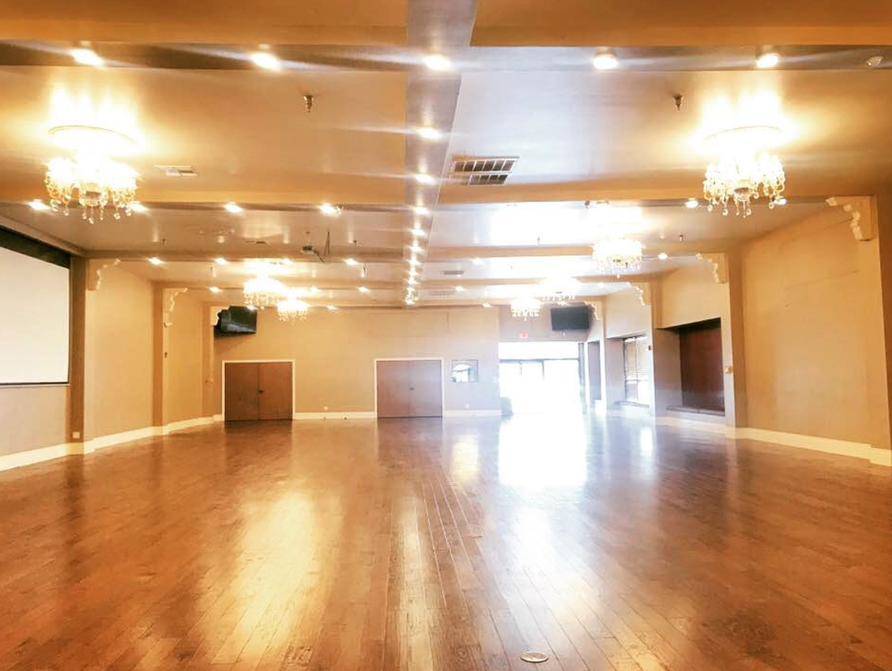 Empty banquet hall with polished wooden floors, chandeliers, and large windows at the back letting in natural light.