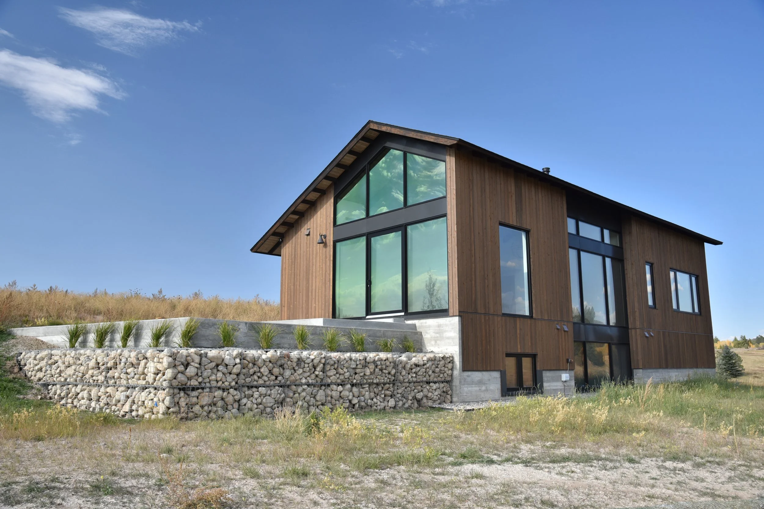 Modern house with wooden exterior, large glass windows, and a sloped roof, situated on a grassy landscape under a blue sky.