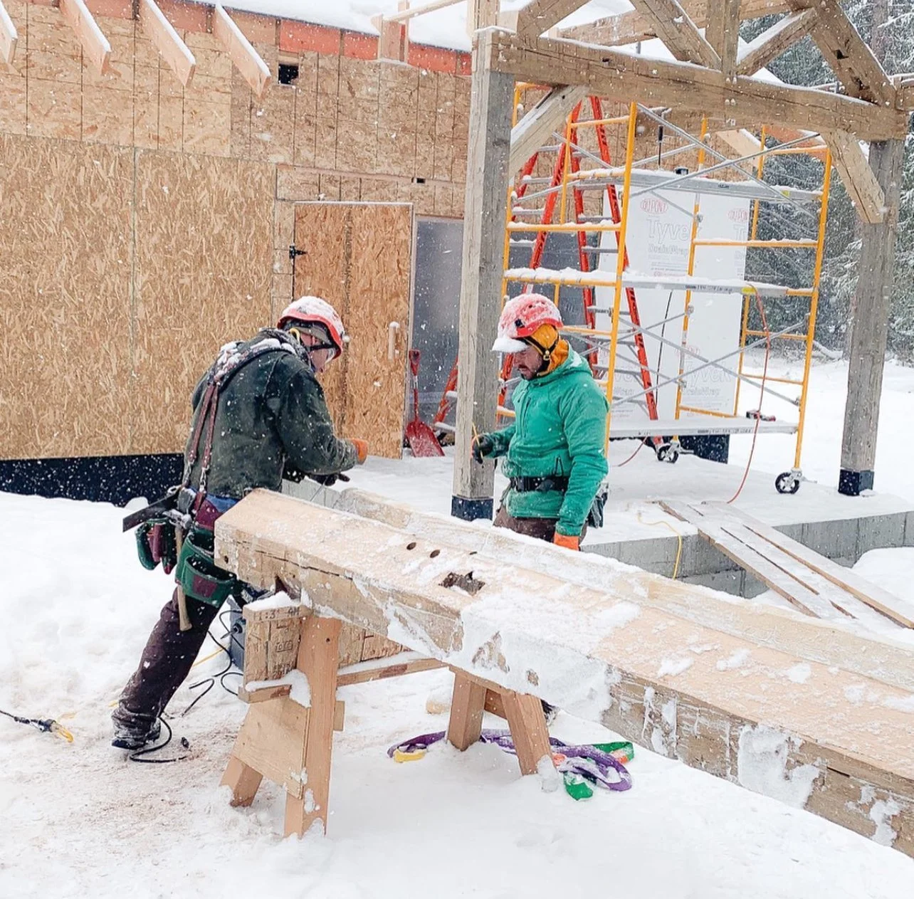 Two construction workers wearing red hard hats and winter gear working outside on a snowy day. They are working on a wooden structure with scaffolding in the background, surrounded by snow.