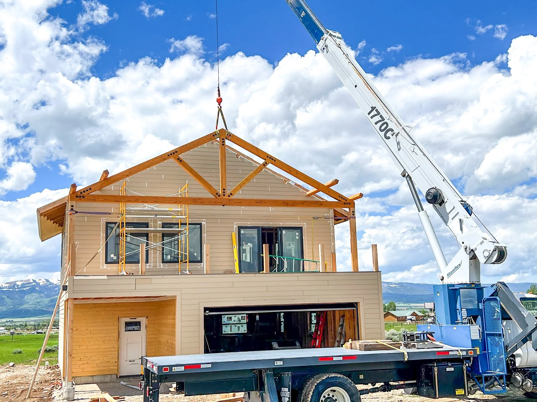 A two-story house under construction with a crane lifting roof trusses into place. The house has beige and wood siding, large windows, and a garage space on the ground floor. Construction materials and equipment are visible around the site.