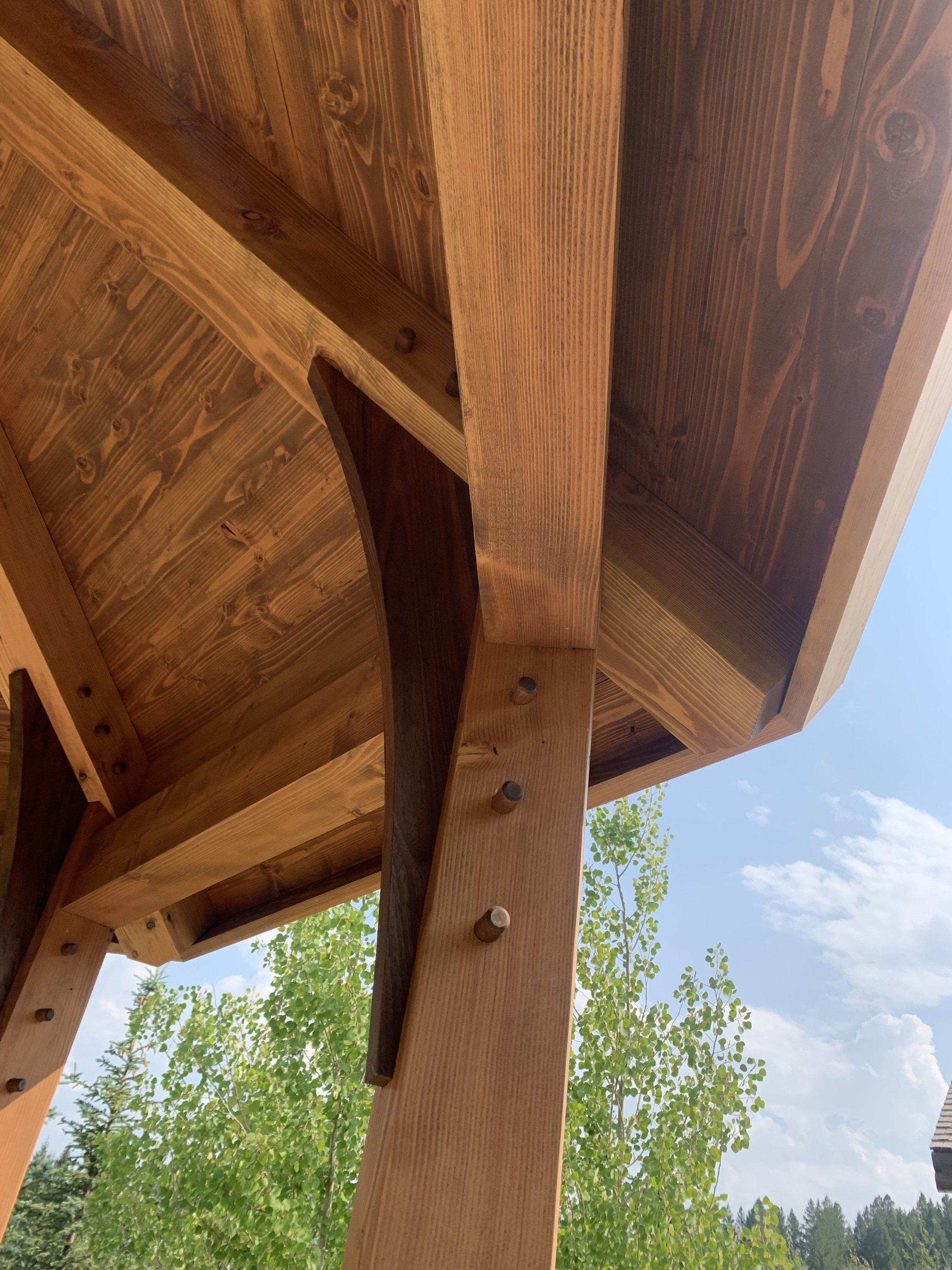 Close-up view of a wooden structure's underside, showing wooden beams and bolts with green trees and a blue sky in the background.