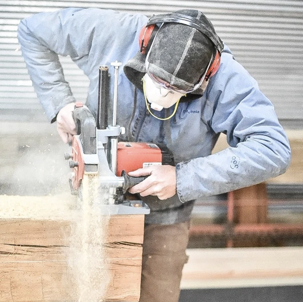 A person wearing safety gear, including a face mask, ear protection, and safety glasses, uses a saw to cut a large wooden log in a workshop.