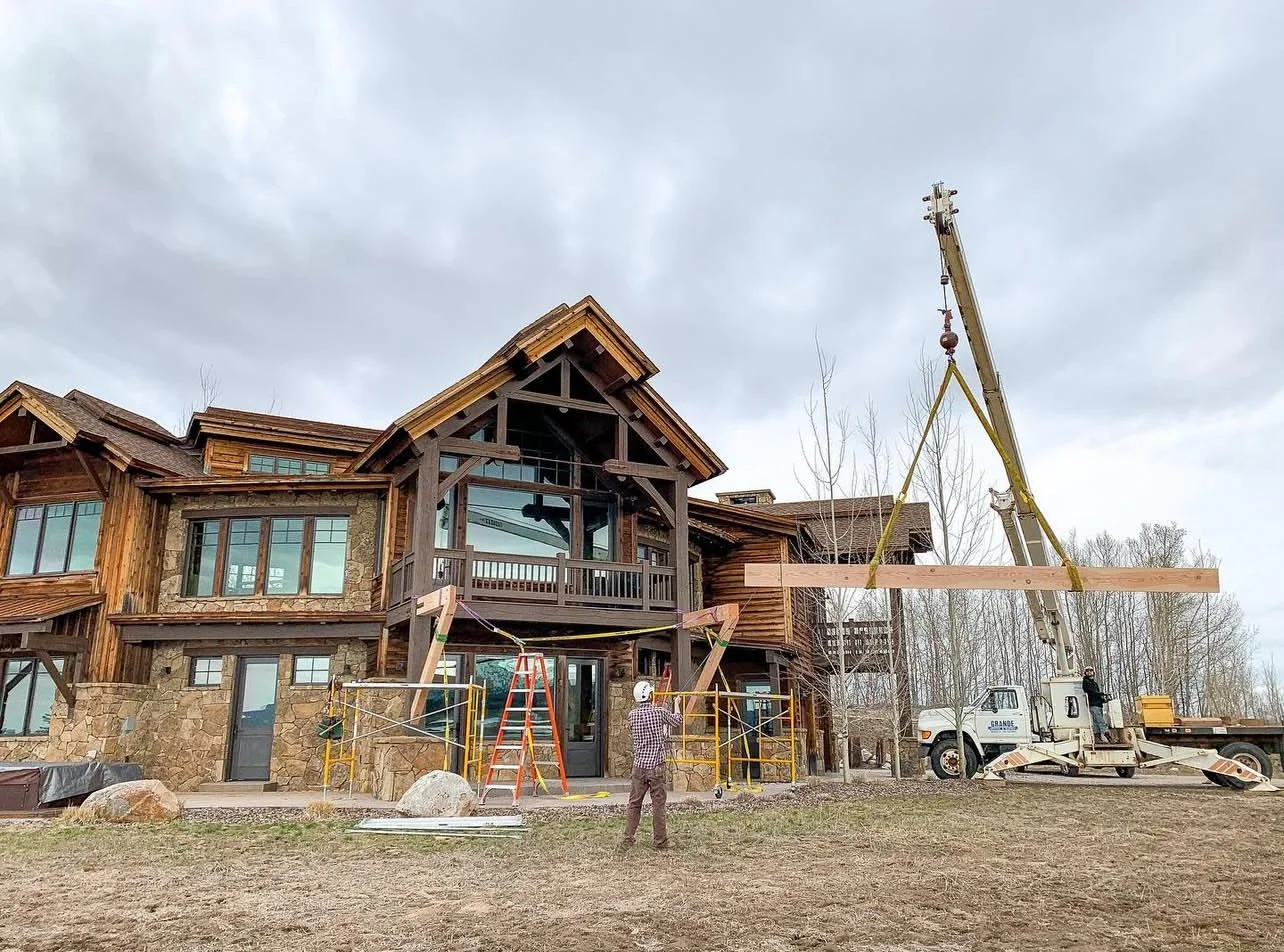 Construction workers are installing a large wooden beam on a multi-story house using a crane, with scaffolding around the house.