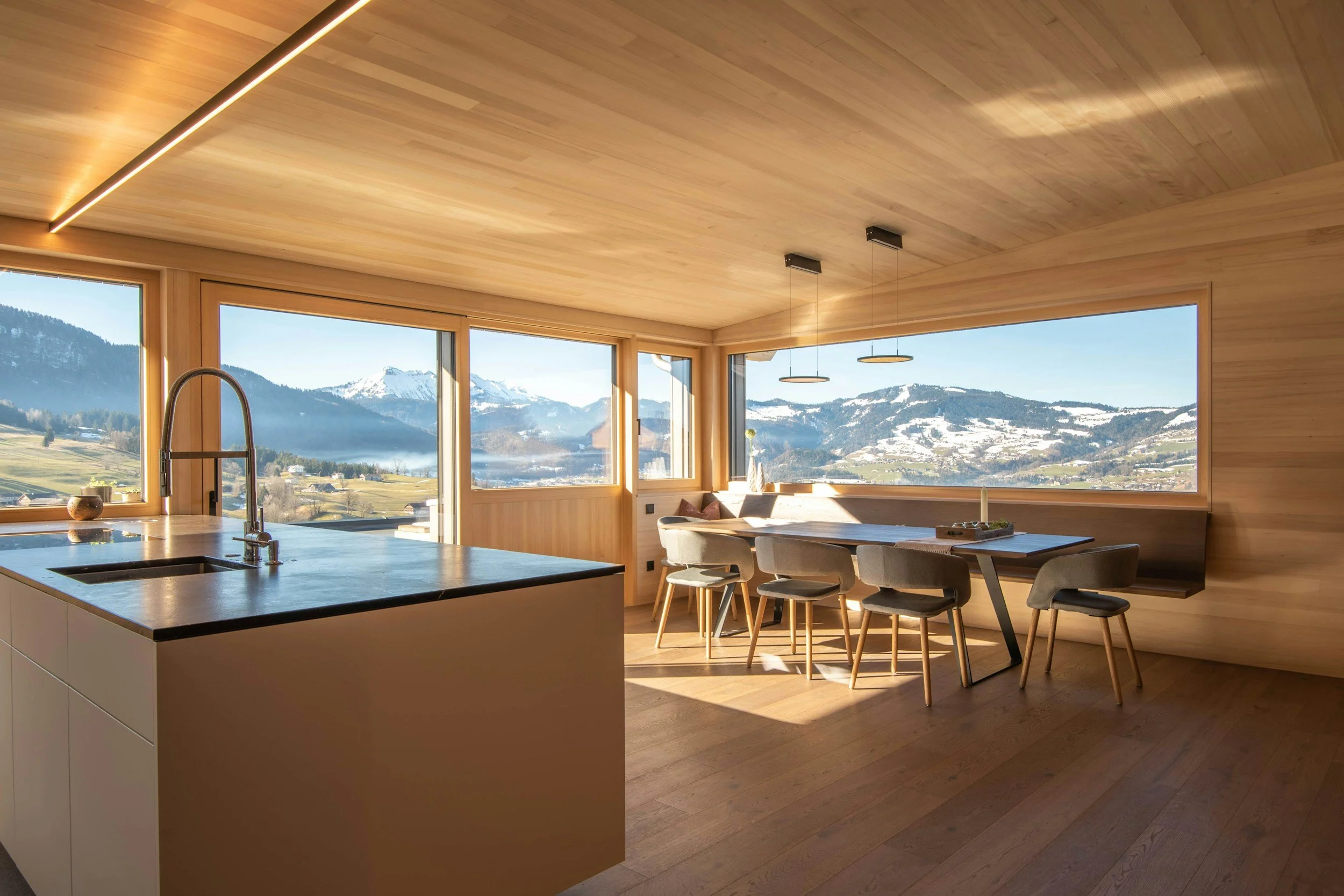 Modern kitchen and dining area with large windows showing mountain landscape, wooden interior, island with sink, dining table with chairs, and minimalist lighting.