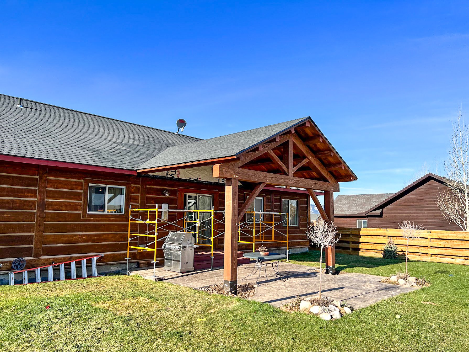 Backyard of a house with a wood deck patio, small trees, a green lawn, and a wooden fence, under a bright blue sky.