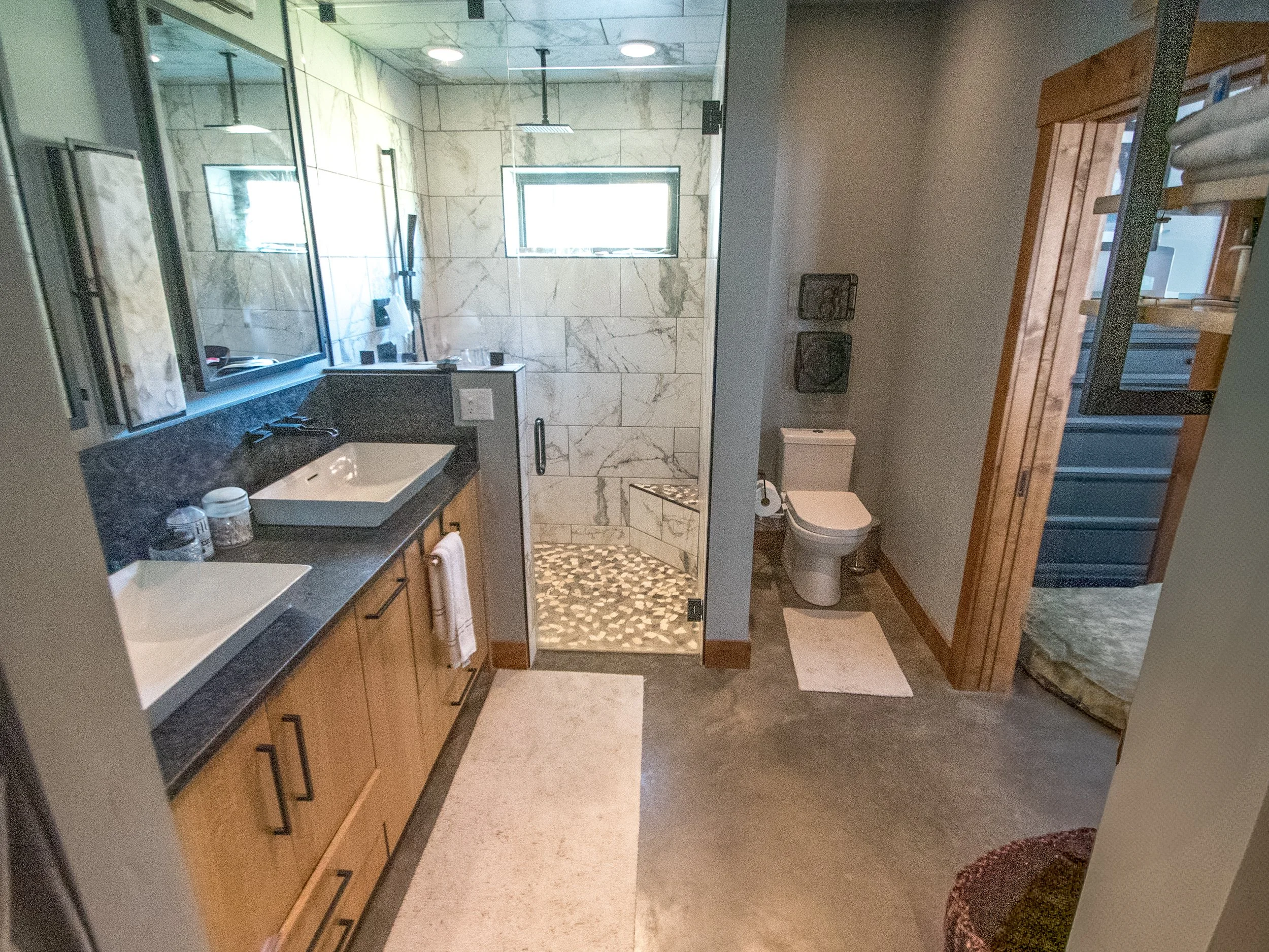 Modern bathroom with double vanity sinks, a glass-enclosed walk-in shower with marble tiles, a small window, a toilet, and a wooden stairway leading upwards.