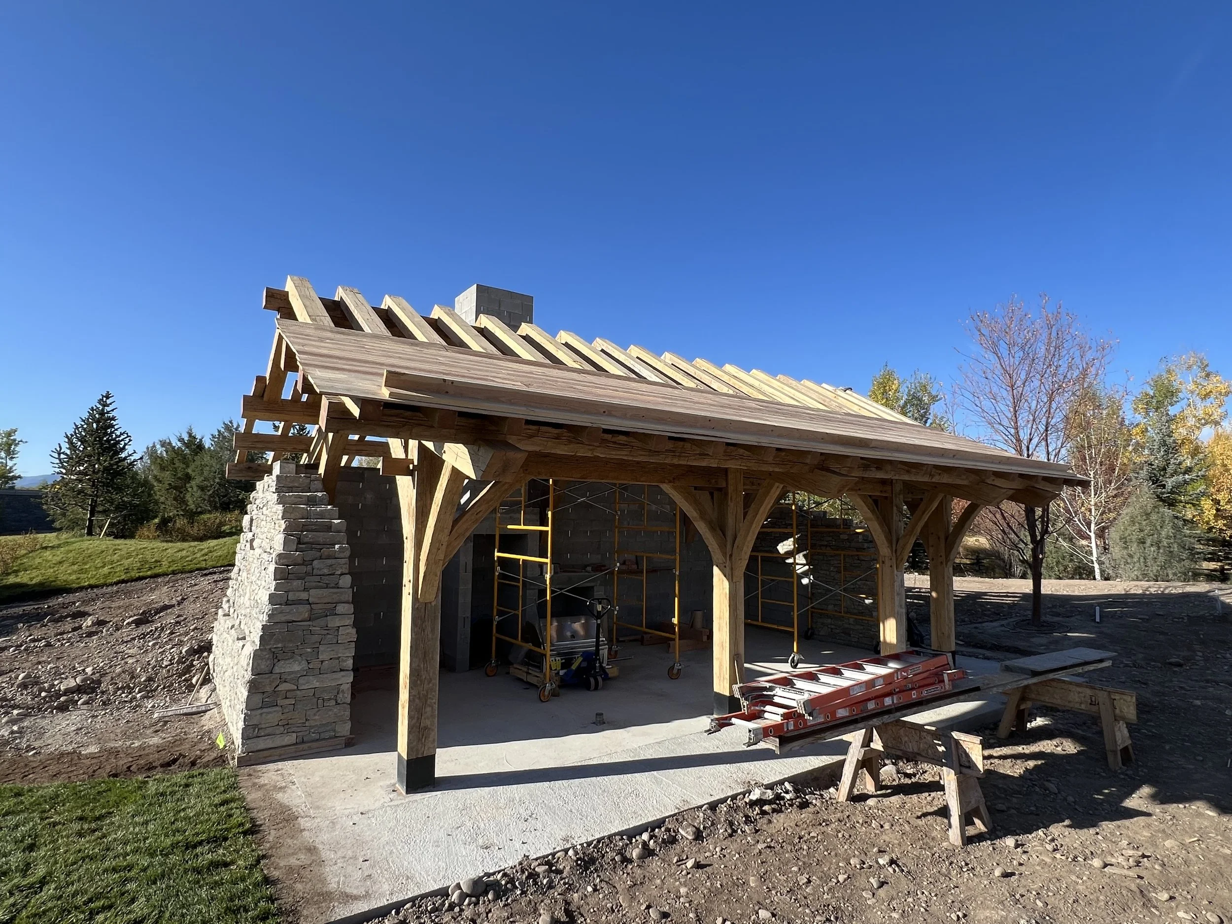 Under-construction wooden structure with stone pillars, scaffolding, and construction tools in a backyard with trees and blue sky.