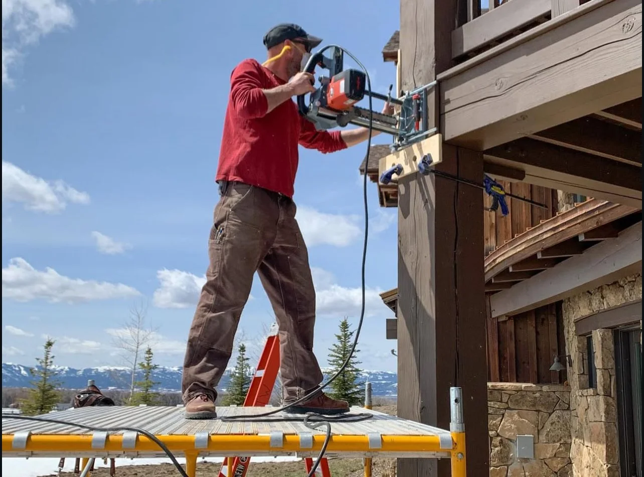 A man wearing a red long-sleeve shirt, brown pants, and a black cap is using a large power tool on the exterior of a wooden building outdoors. He is standing on a metal scaffold with a partly cloudy sky and distant mountains in the background.