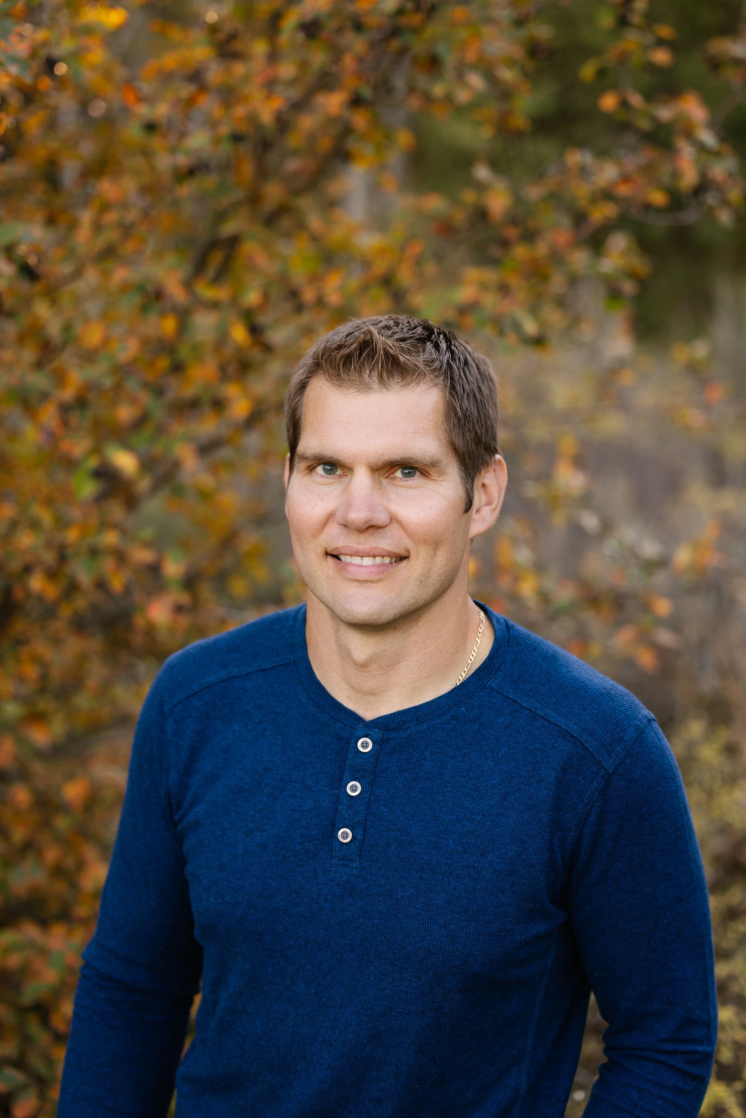 A young man with short brown hair and blue eyes wearing a dark blue long-sleeve shirt with buttons, standing outdoors in front of autumn-colored trees.