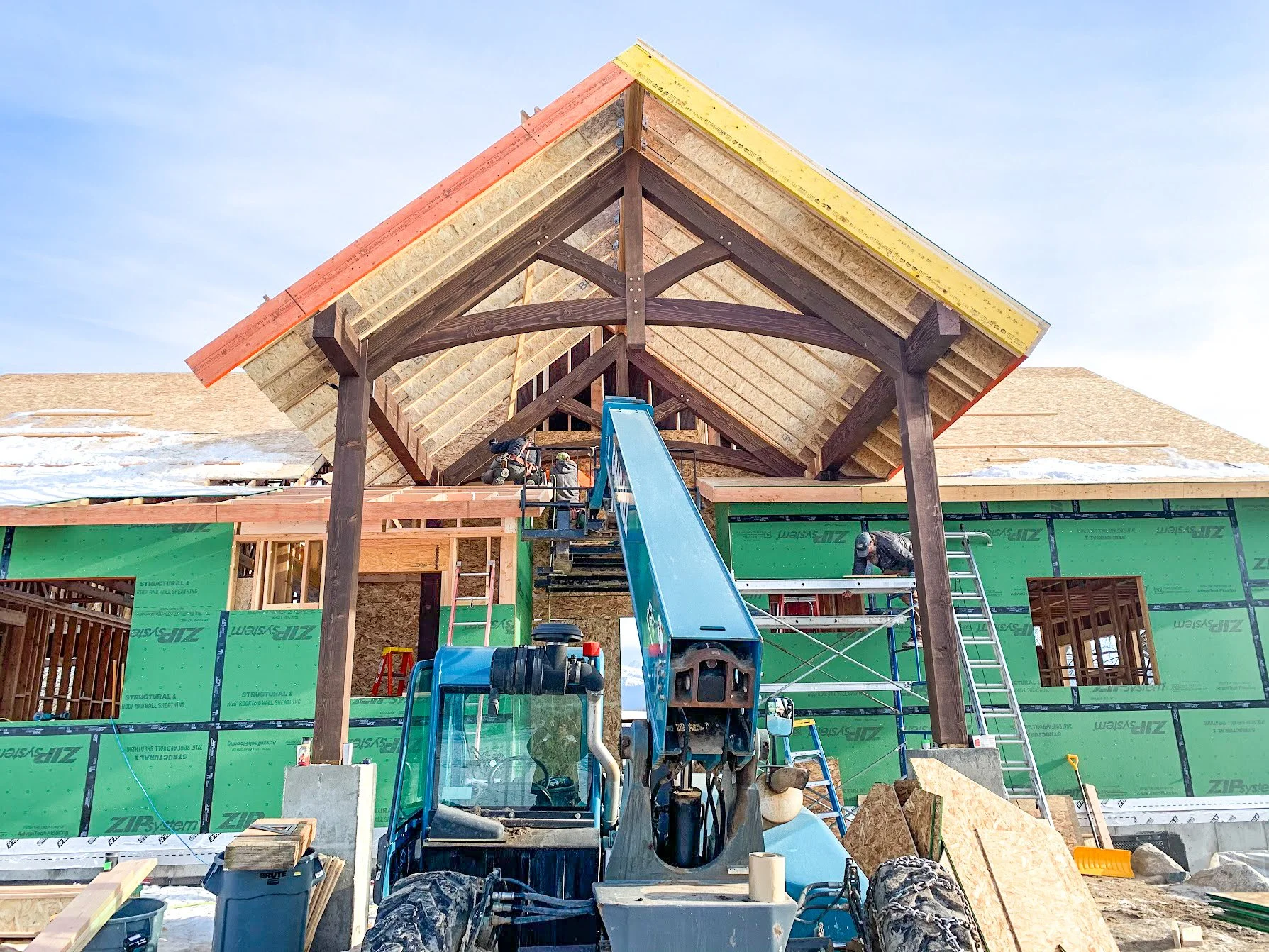Construction site of a house with workers, a lift, and scaffolding, showing the roof framing and partially installed roof.