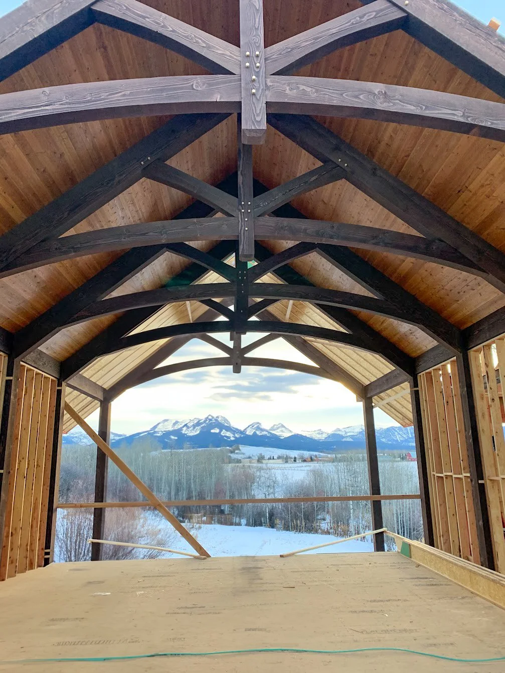 Interior view of a partially constructed building with exposed wooden framing and trusses, overlooking snow-covered landscape with mountains in the distance.