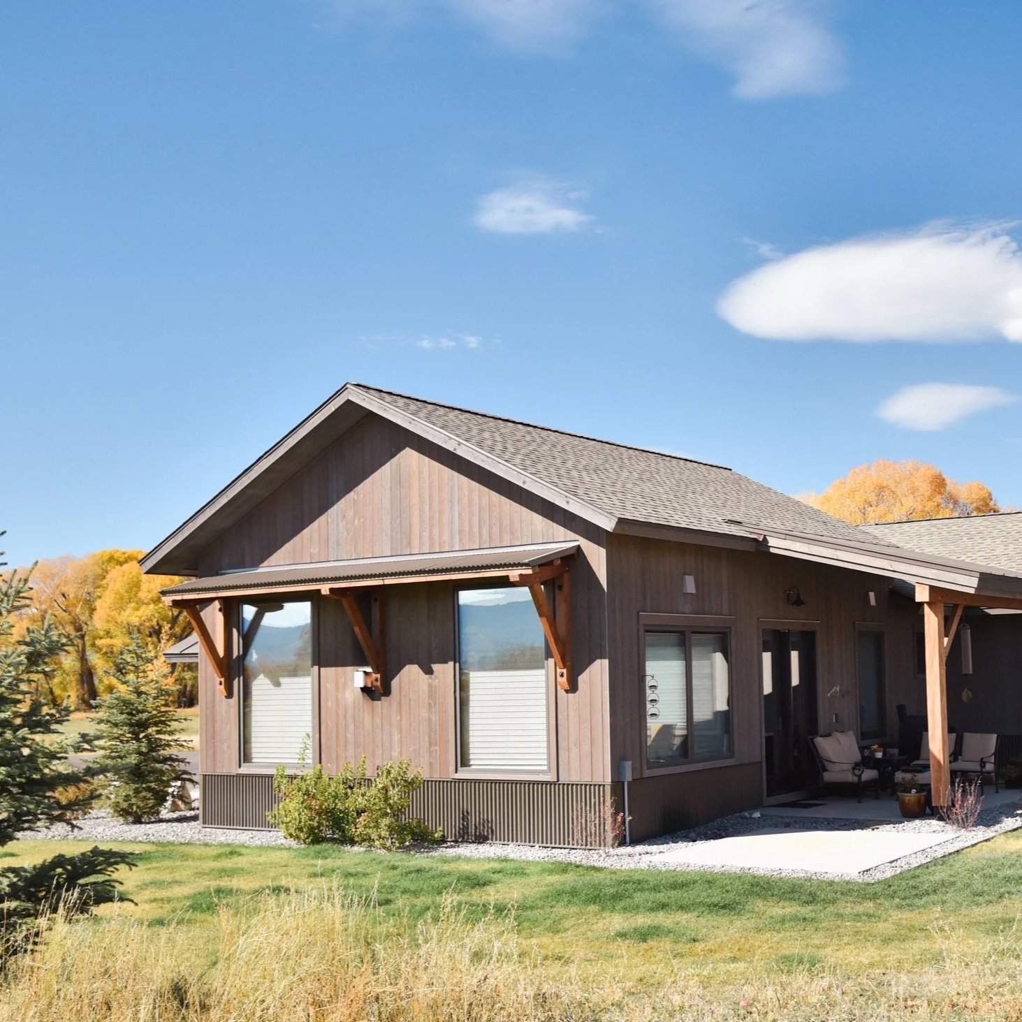 A house with wooden siding, a sloped roof, and a covered porch with outdoor furniture, set against a background of trees with fall foliage and a blue sky with white clouds.