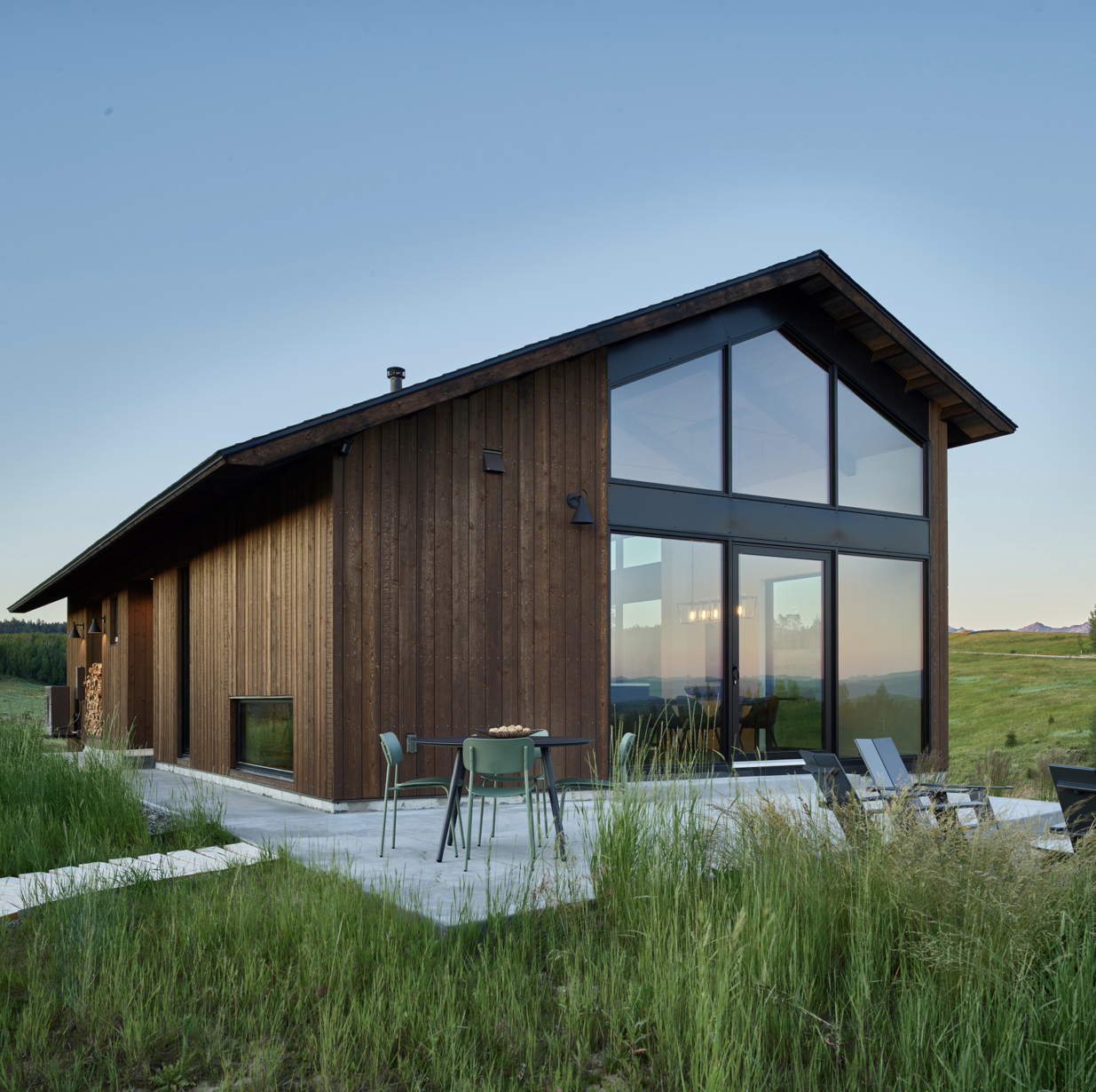 Modern house with wooden exterior and large glass windows featuring a patio with outdoor furniture and green surroundings at sunset.