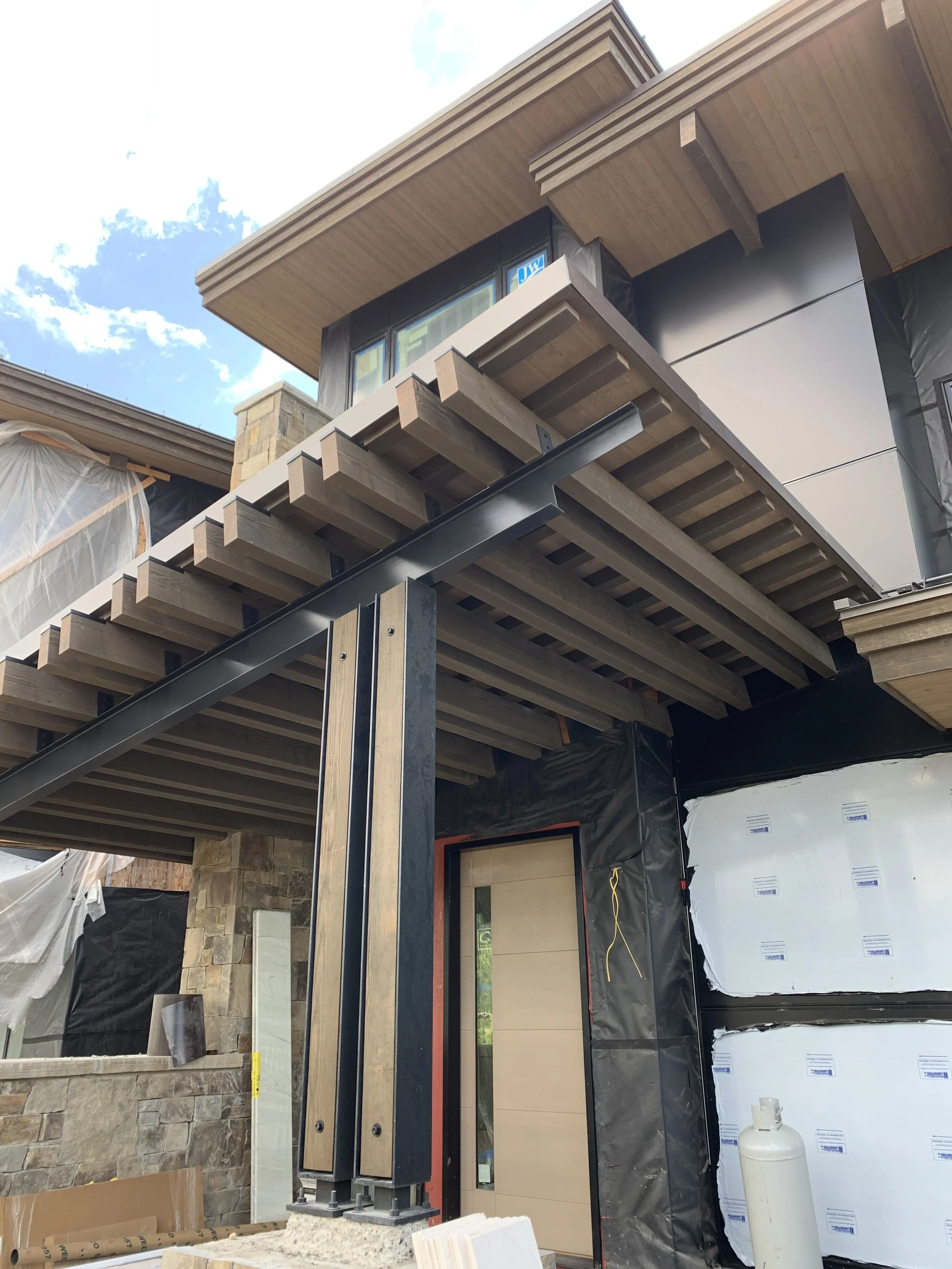 Under construction modern house with wooden balcony, stone wall, and black metal support pillar, with construction materials and unfinished walls visible.