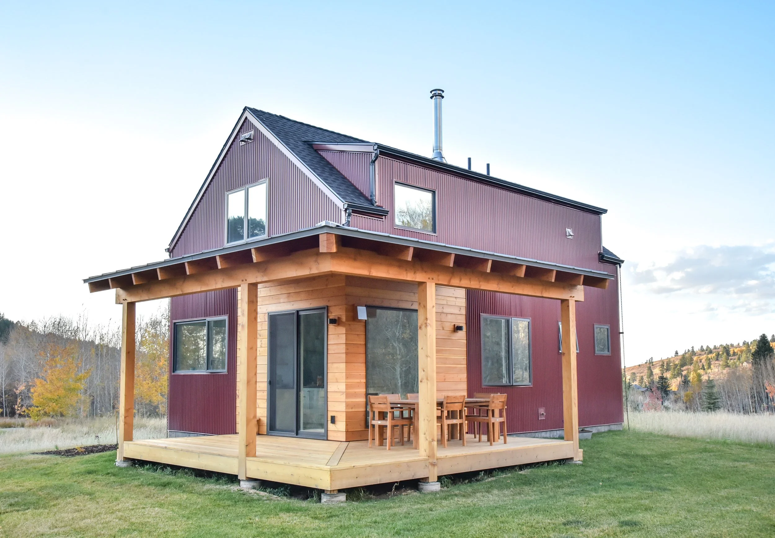A two-story house with a red metal exterior and a new wooden deck with outdoor furniture, set in a grassy yard with trees in the background.