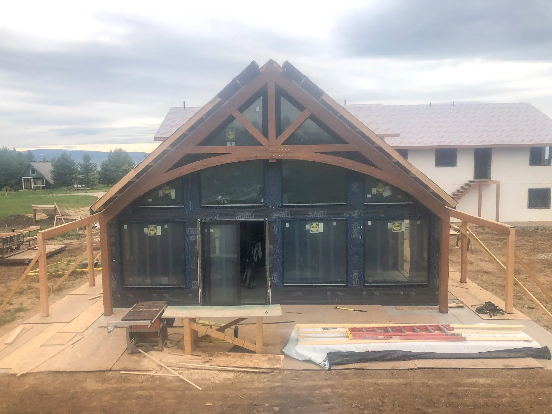 Construction site of a house with a prominent A-frame front, large glass windows, and wooden framework, with another house in the background and a cloudy sky above.