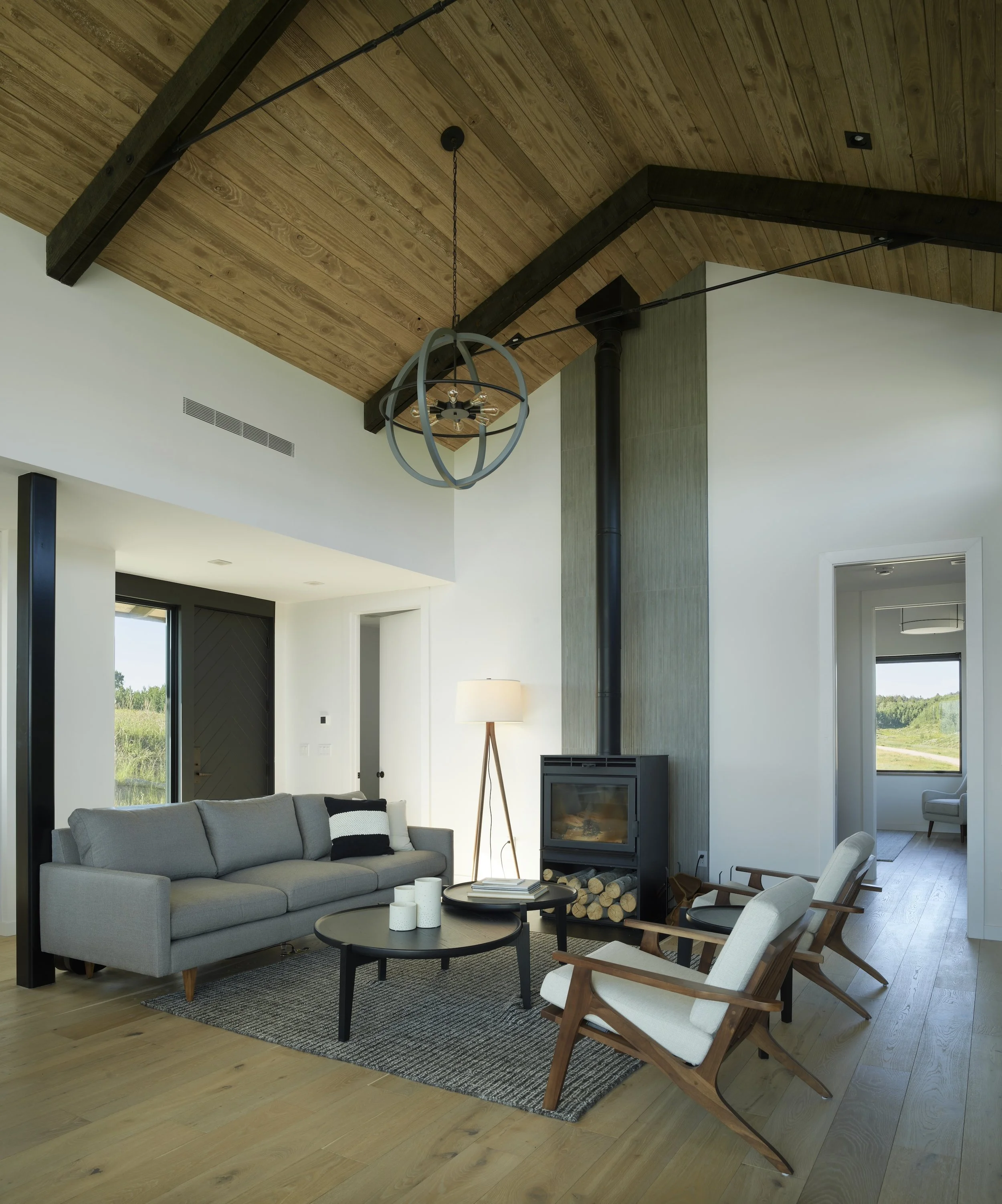 Living room with gray sofa, wooden chairs, fireplace, wood ceiling, black and gray decor, large window with grass view.