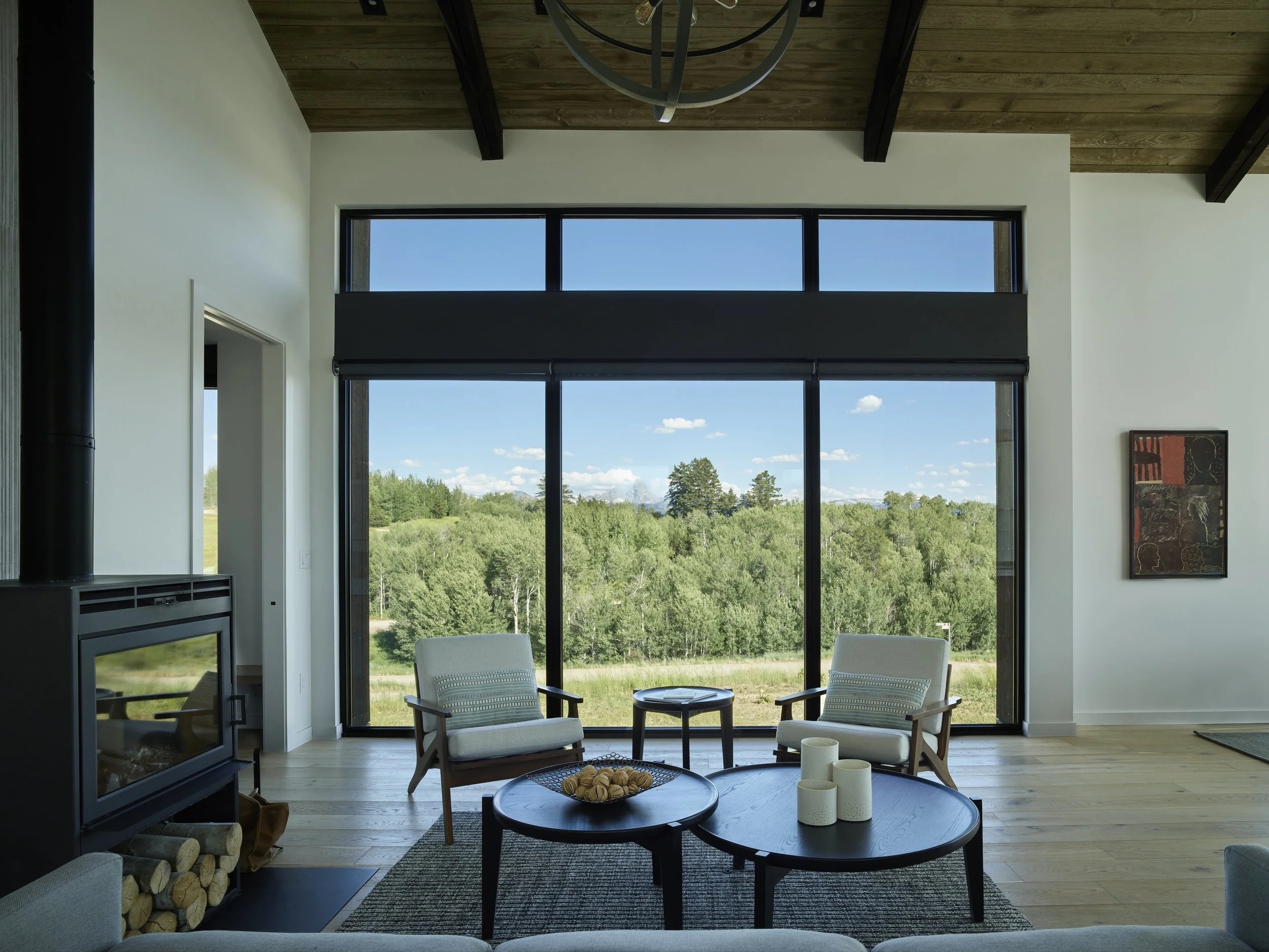 Living room with large floor-to-ceiling windows showing green trees outside, two armchairs, a round black coffee table with candles, a smaller black side table, and a wood stove, with a wooden ceiling and modern decor.