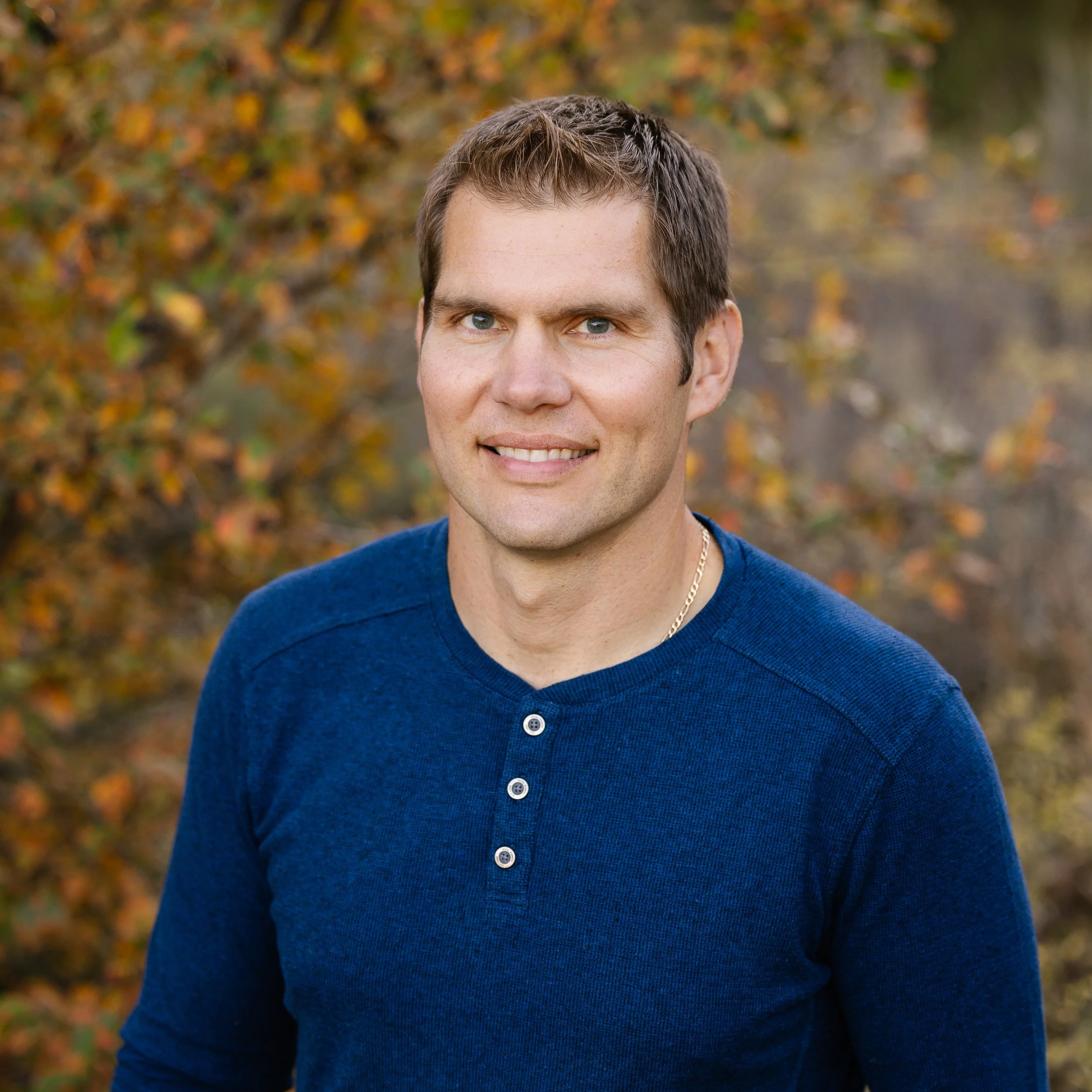 A man with short brown hair and blue eyes smiling outdoors surrounded by autumn foliage, wearing a blue long-sleeve shirt and a gold chain necklace.