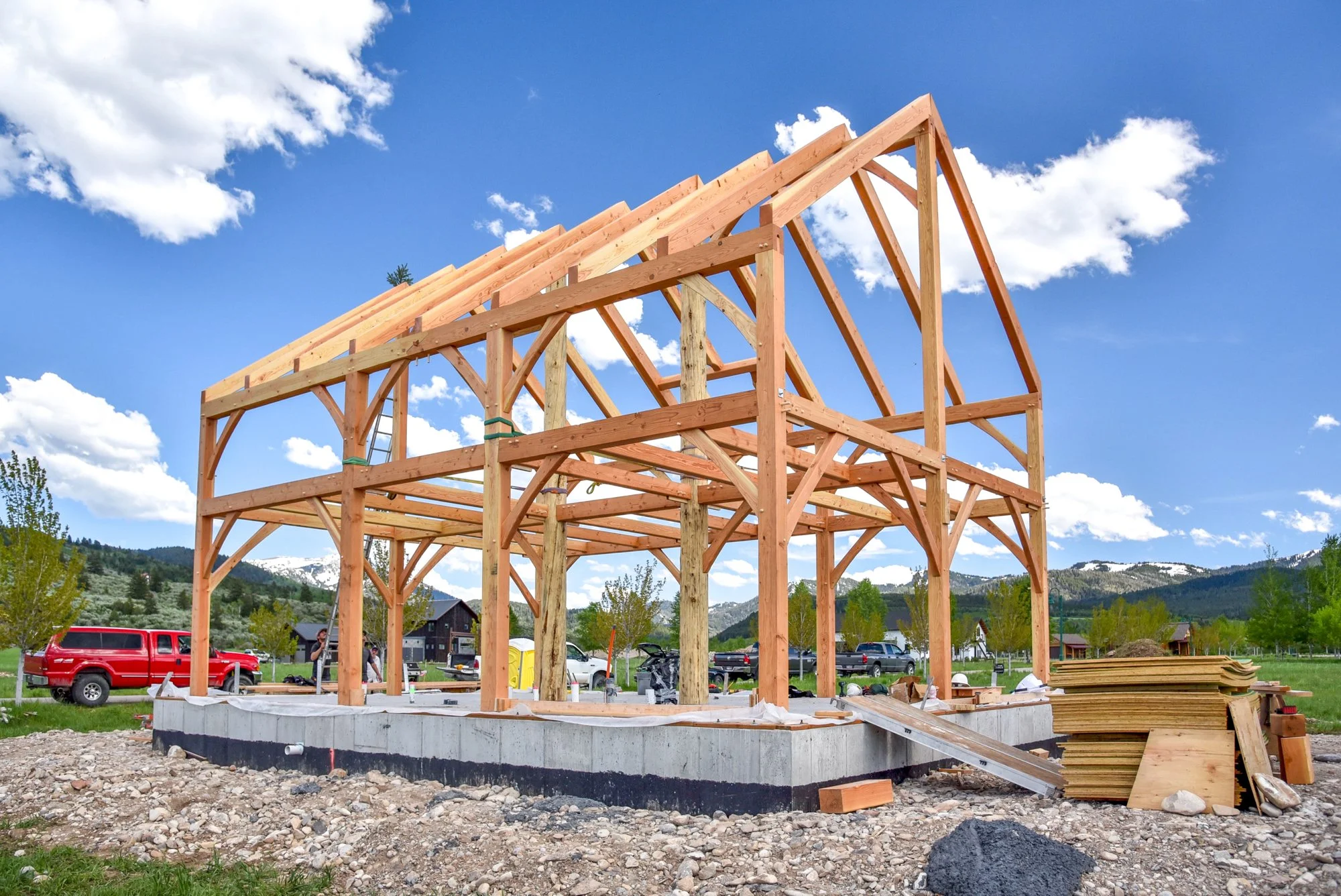 A house under construction with a wooden framework on a concrete foundation, set against a scenic backdrop of mountains, trees, and a partly cloudy sky.