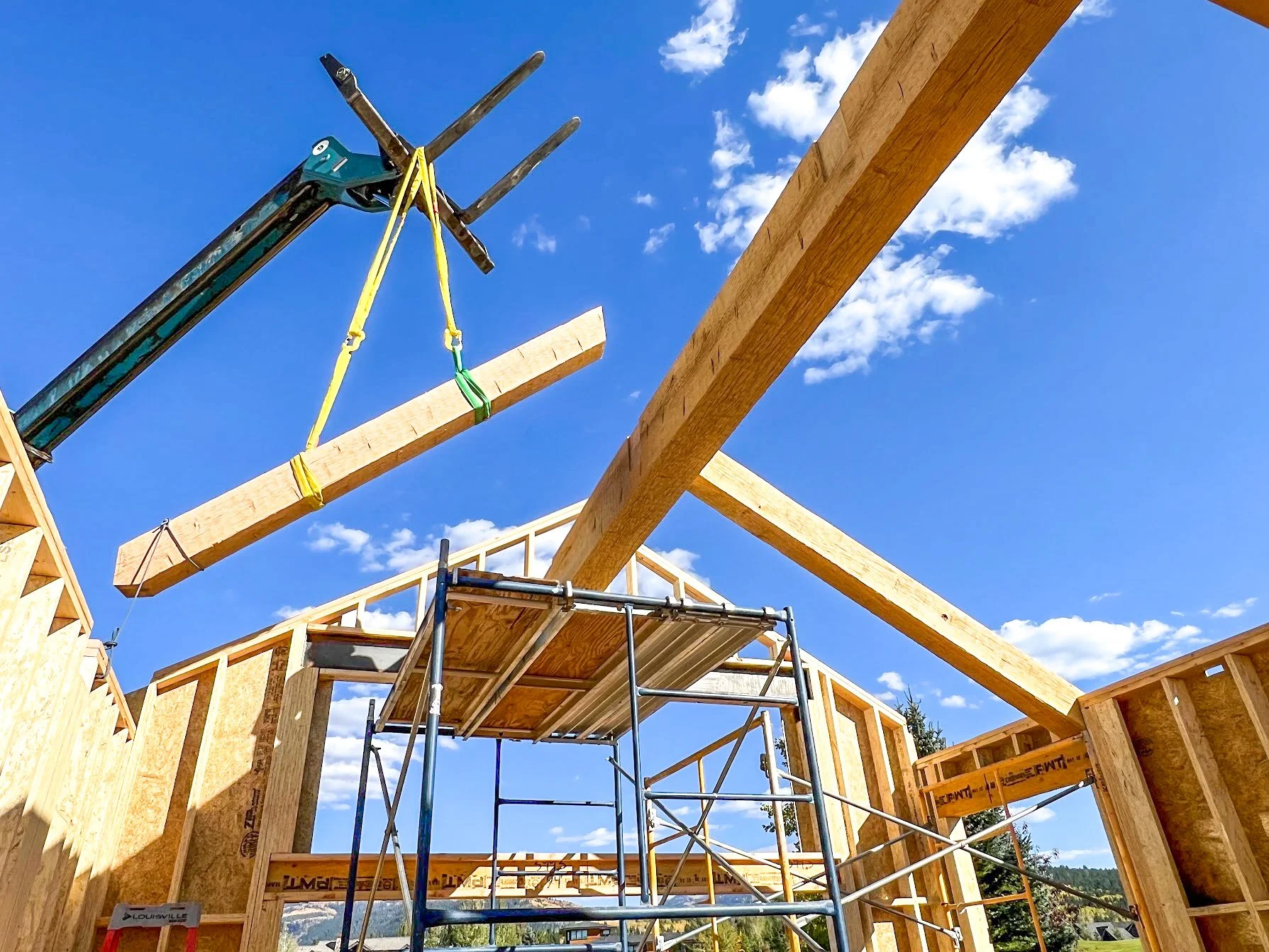 Wooden framing for a building under construction with a crane lifting a long wooden beam into place on a bright, partly cloudy sky background.