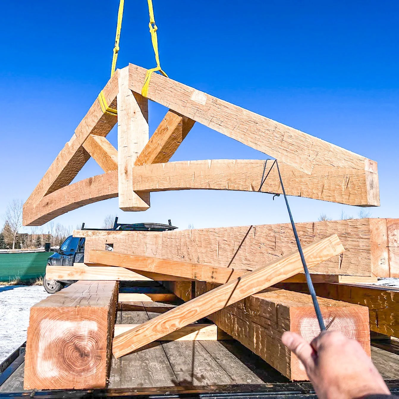 A person holding a black cable, lifting a large wooden framework with a crane. The framework consists of massive wooden beams, with a clear blue sky and some trees in the background.