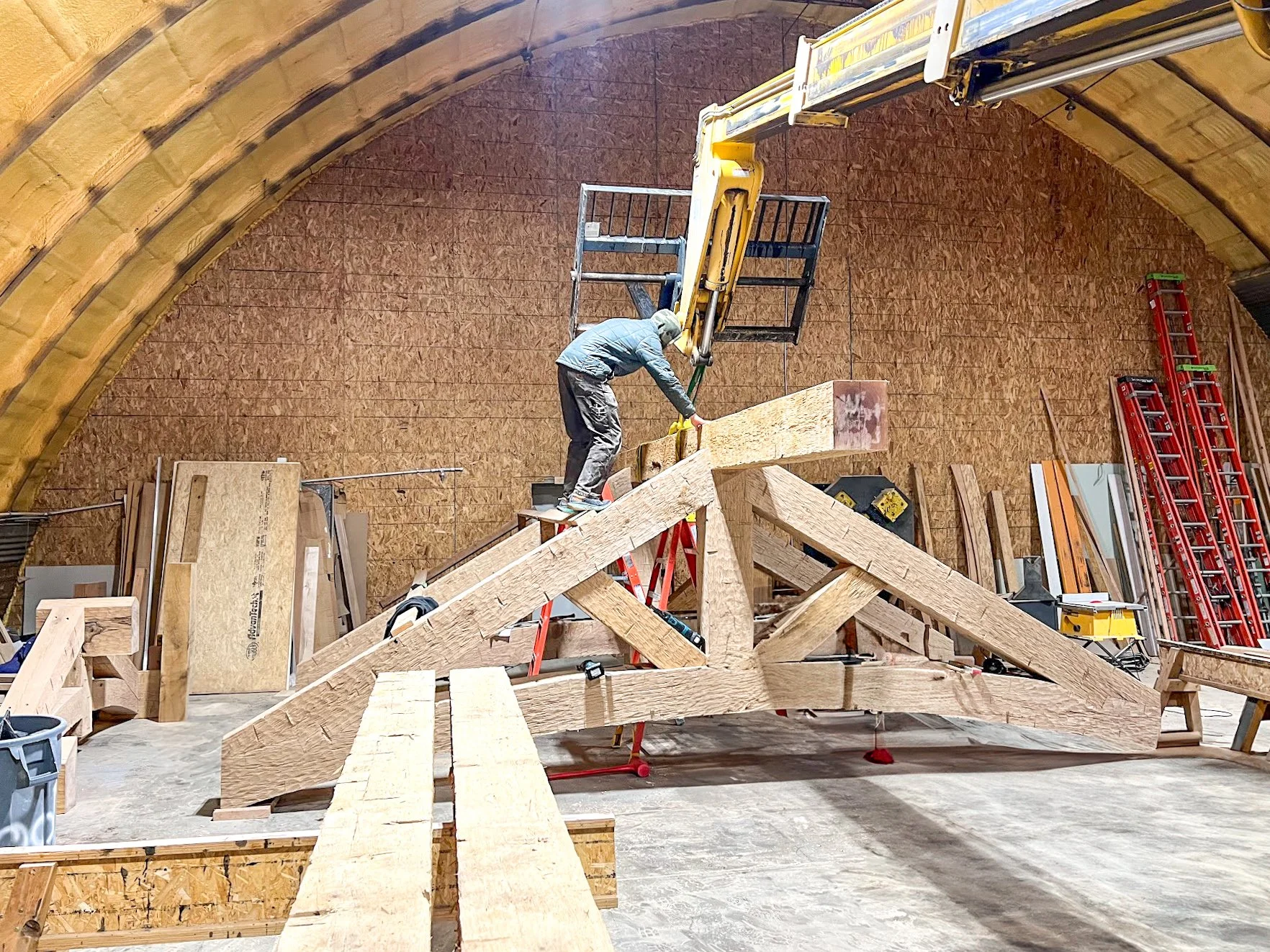 A person working on a construction site inside a large arched wooden structure, standing on scaffolding and working on a large wooden framework.