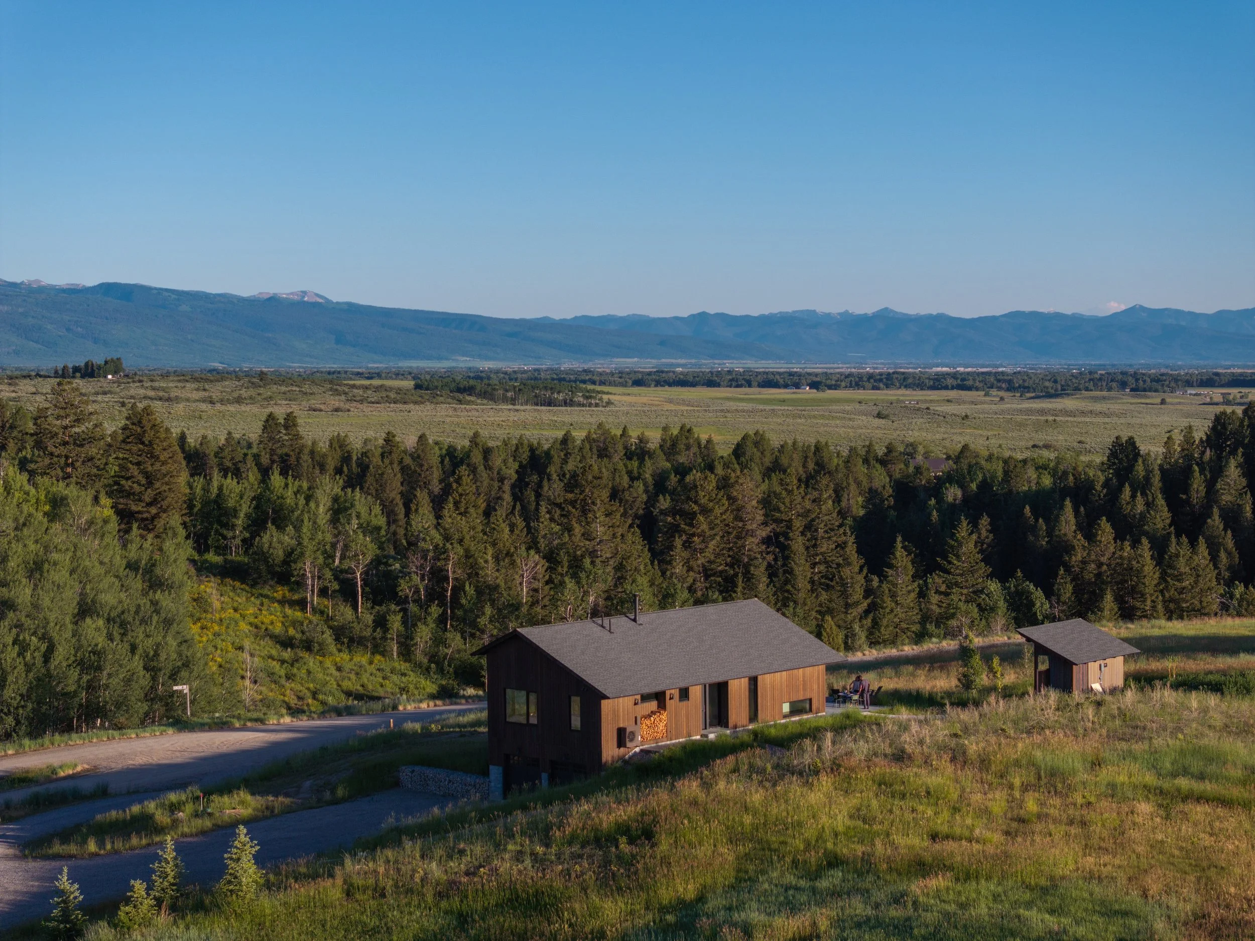 A house located on a hilltop with mountains in the background, surrounded by trees and open fields under a clear blue sky.