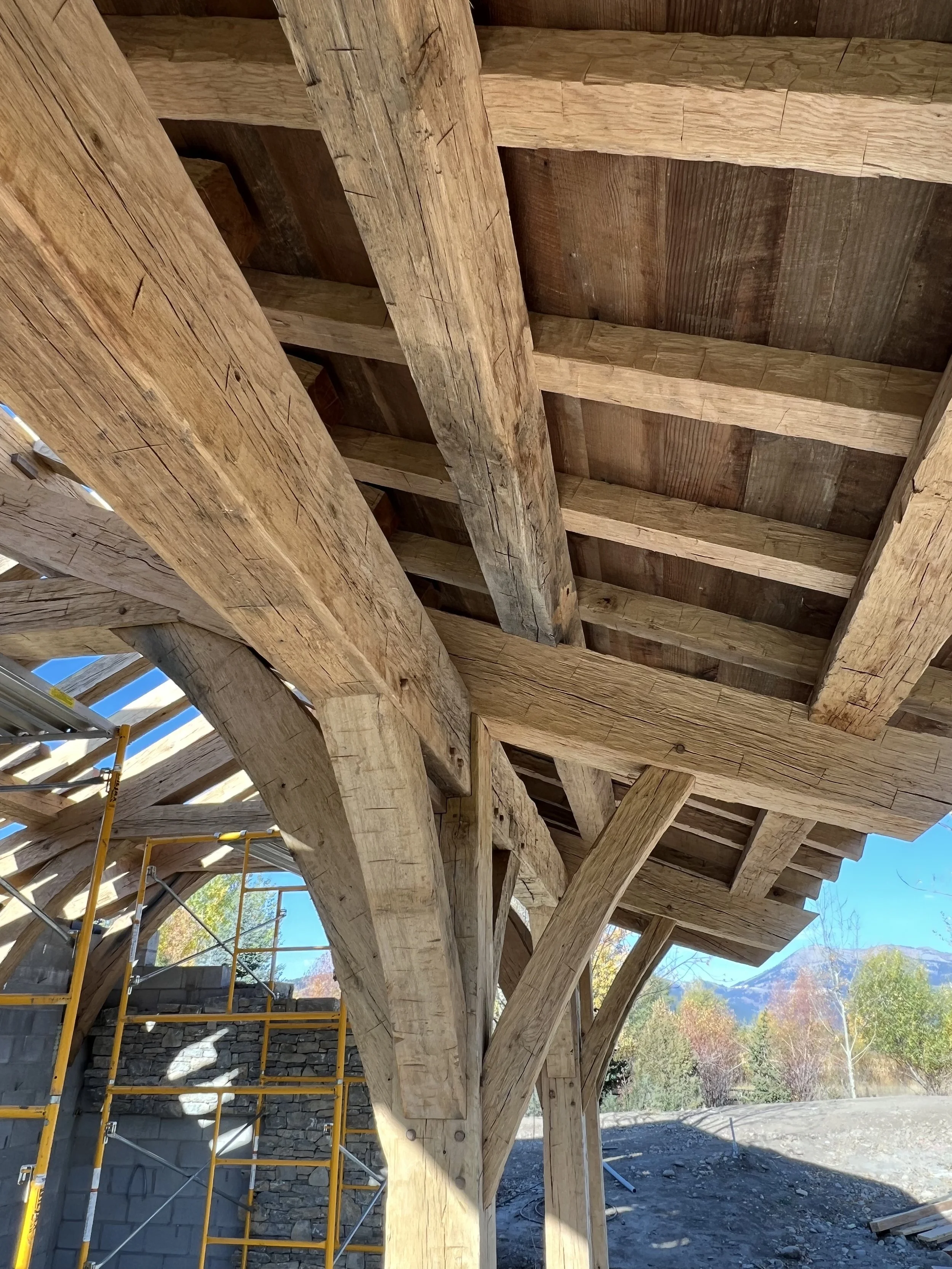 Close-up view of a wooden roof structure with large wooden beams and supports, with a blue sky and distant trees and mountains in the background.