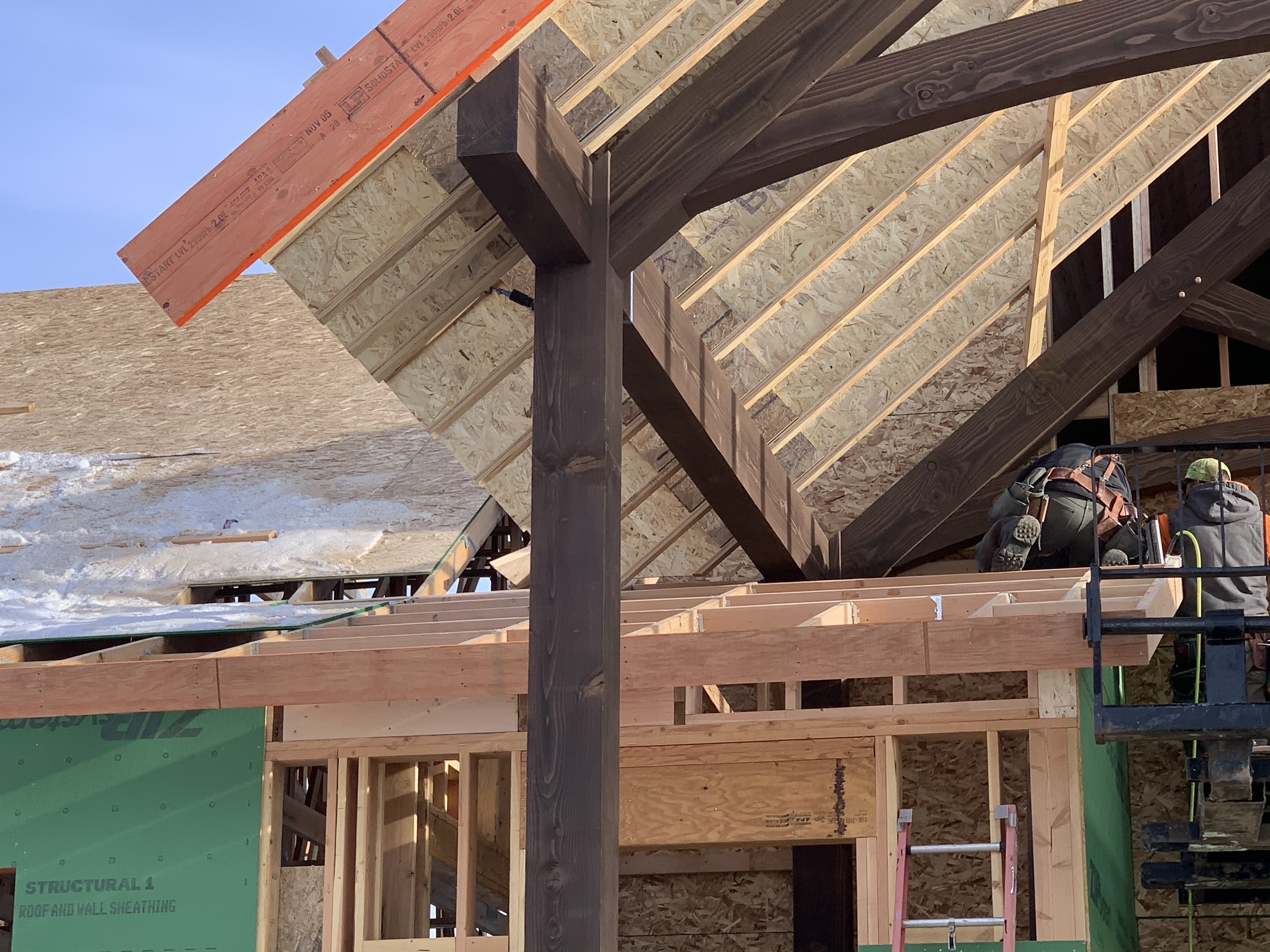 Construction site with workers installing roofing on a wooden structure. The roof is partially covered with sheathing and the workers are on a lift, working near the roof edge. Some snow is visible on the ground.