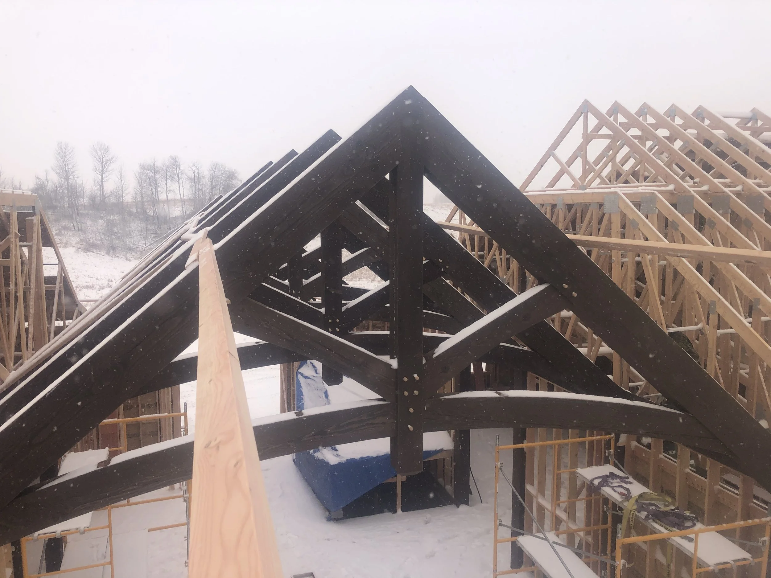 Construction site of a wooden house framing with snow on the beams, snowy landscape in the background.