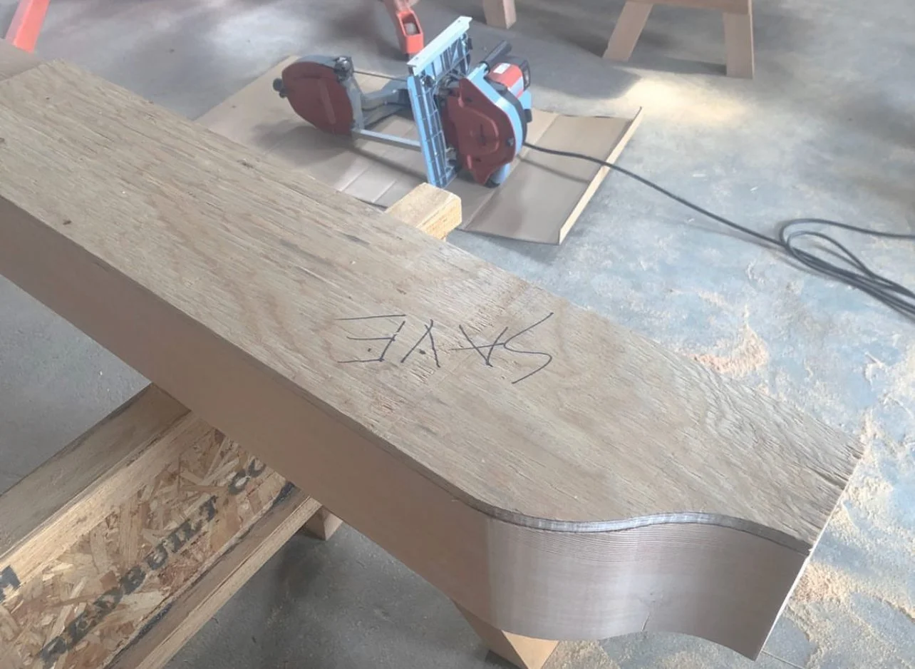 A woodworking workshop with a piece of wood on a workbench, marked with the word 'STYLE' written in black marker. In the background, there is a circular saw on the floor and other woodworking tools and materials.