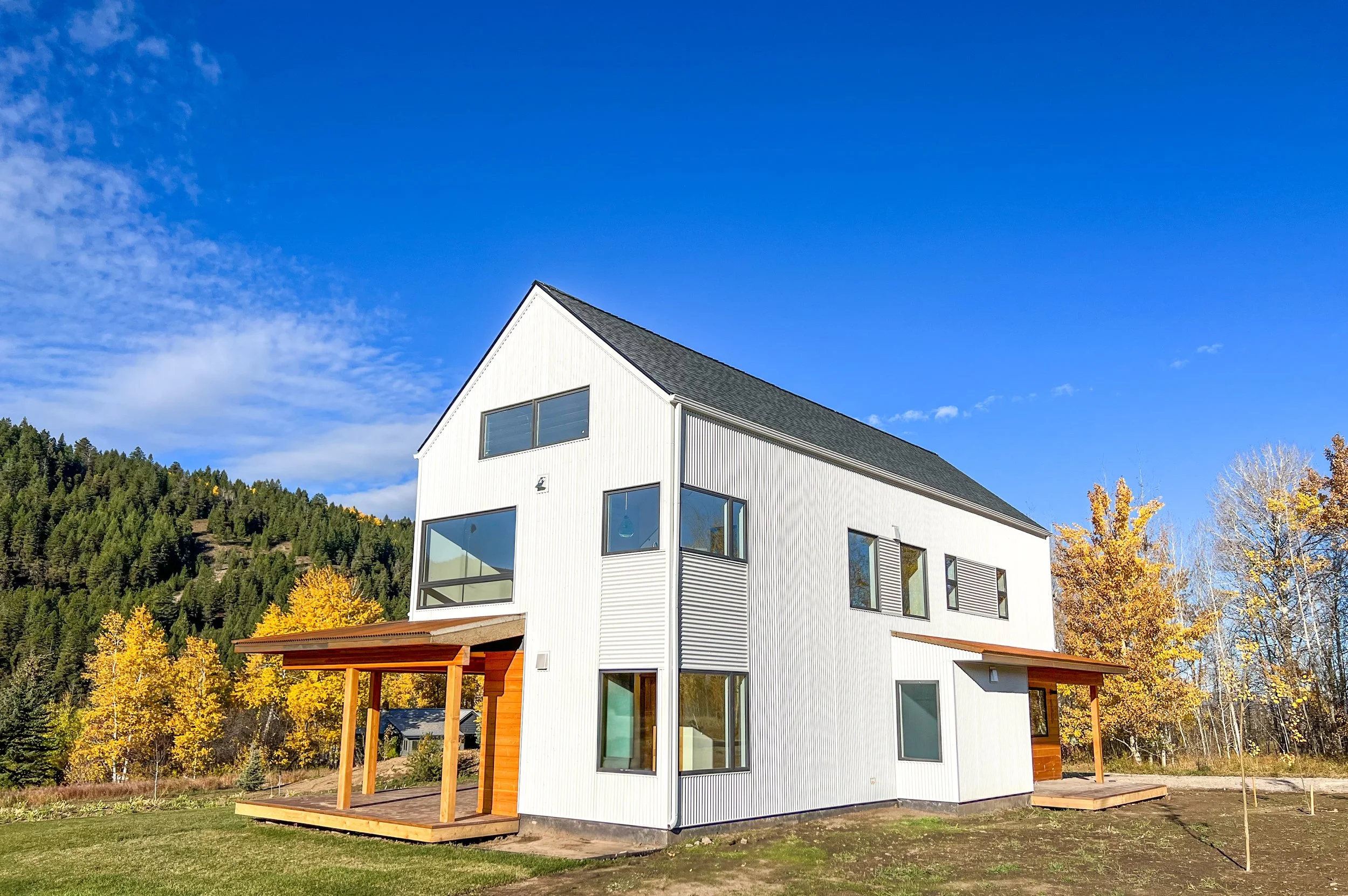 Modern white house with wooden porches in a scenic landscape with trees and hills in the background under a clear blue sky.