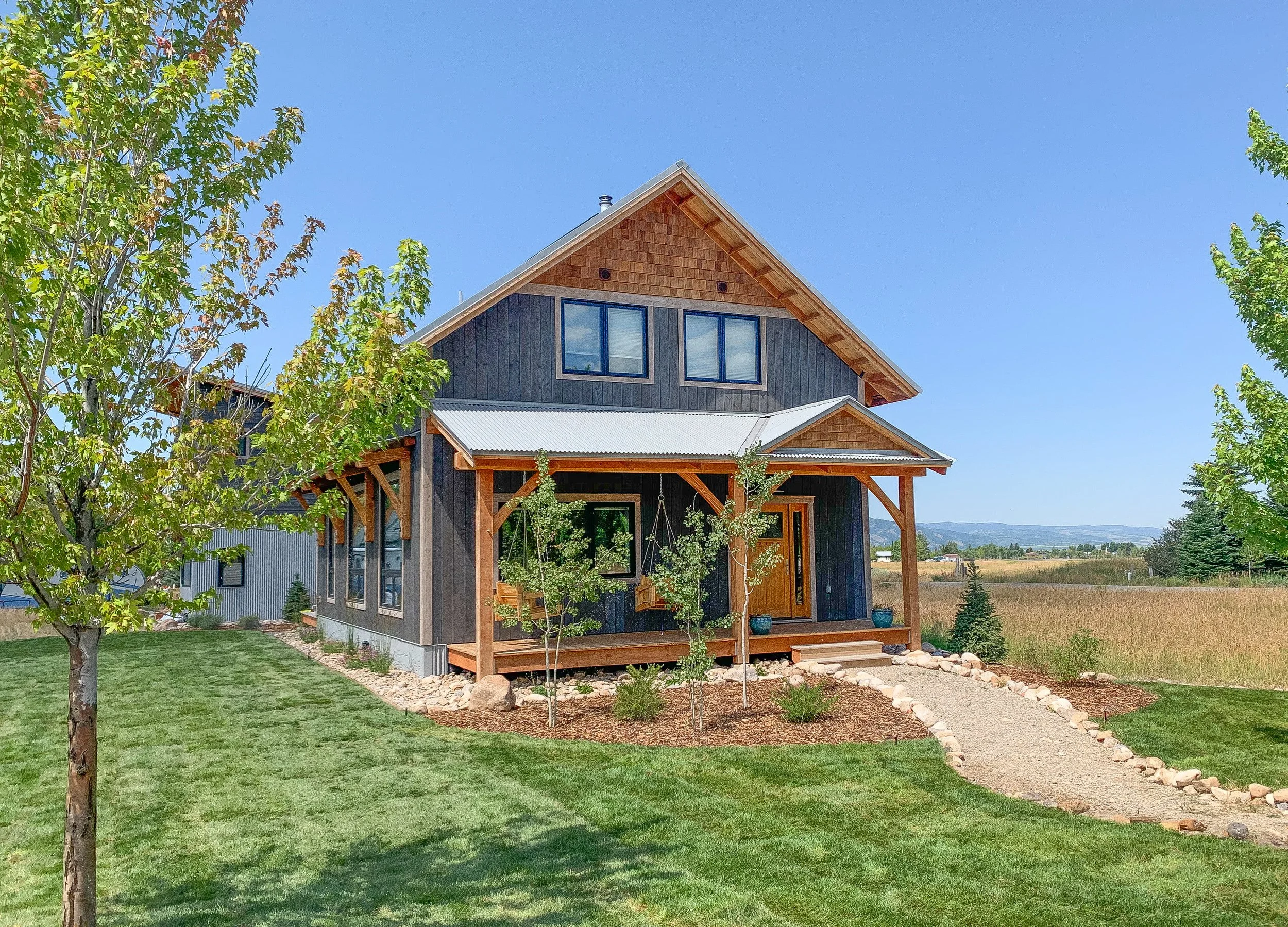 A two-story house with dark wood siding, a metal roof, and a small front porch with swings, surrounded by a manicured lawn and trees, with open fields and blue sky in the background.