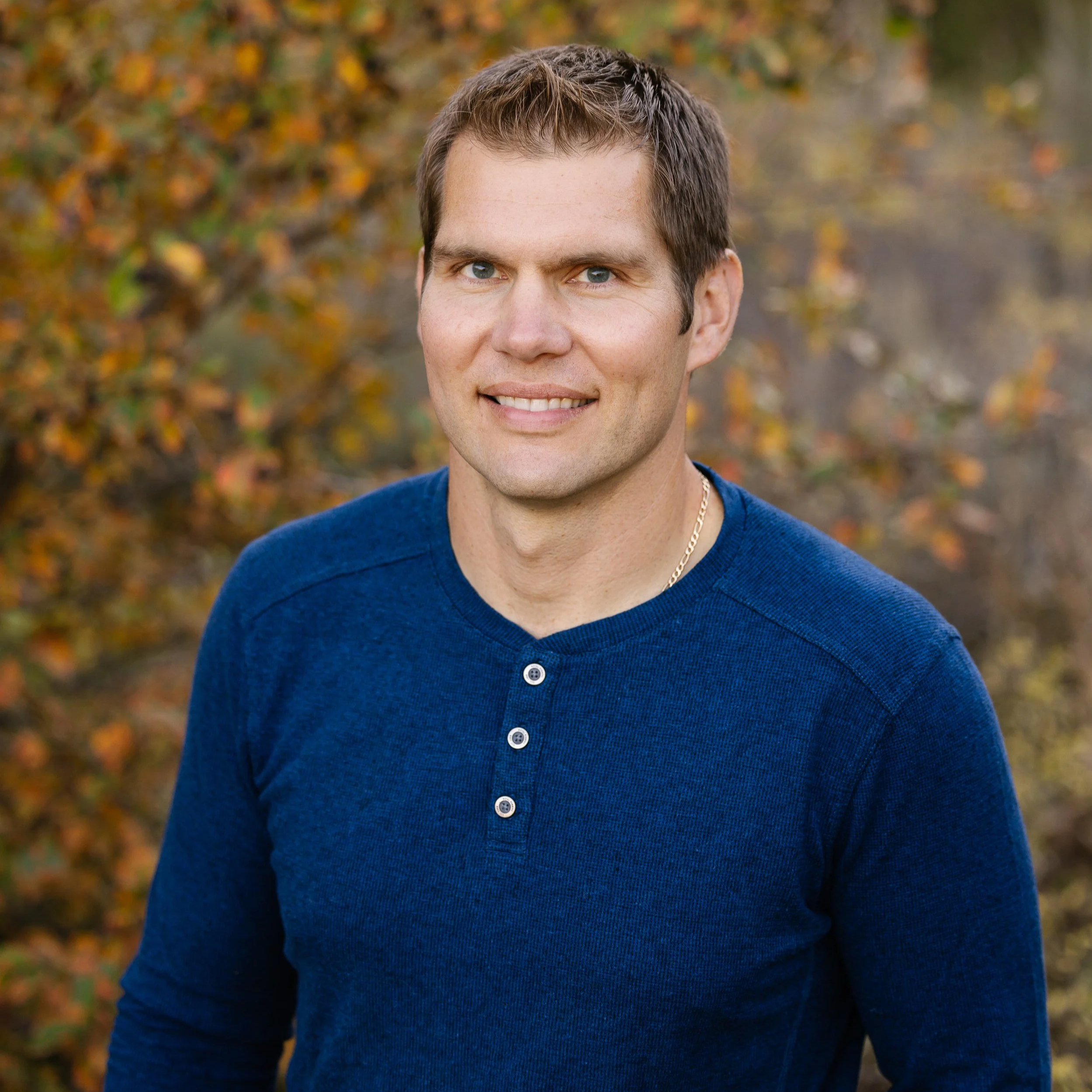 A man with short brown hair, blue eyes, and light skin wearing a navy blue long-sleeved shirt with buttons and a gold chain necklace, standing outdoors with trees and autumn leaves in the background.