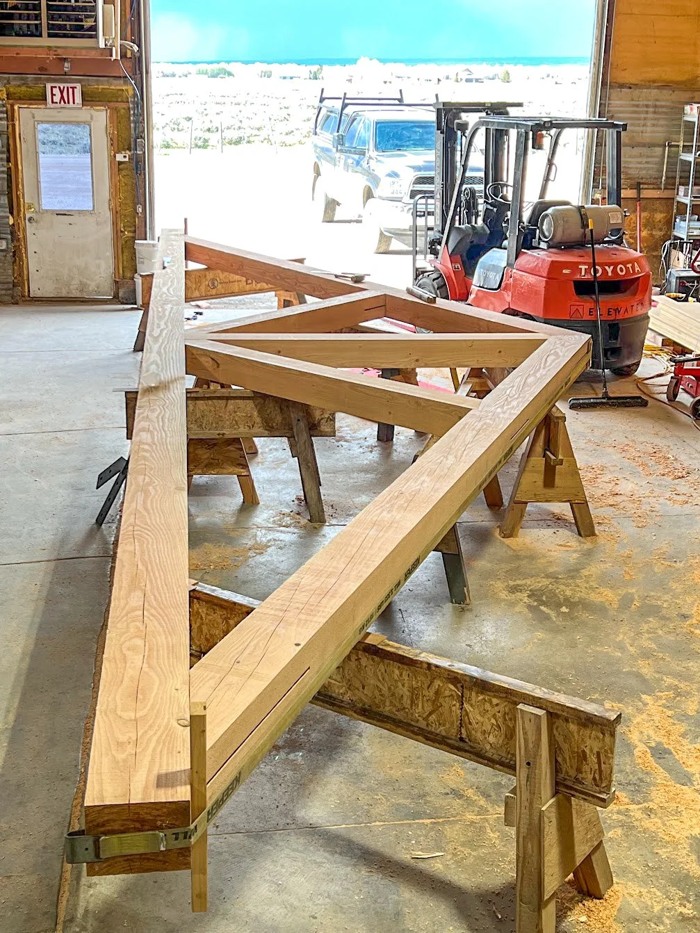 Woodworking workspace inside a workshop with a large wooden frame on workbenches, a red Toyota forklift, a pickup truck outside, and tools on shelves.