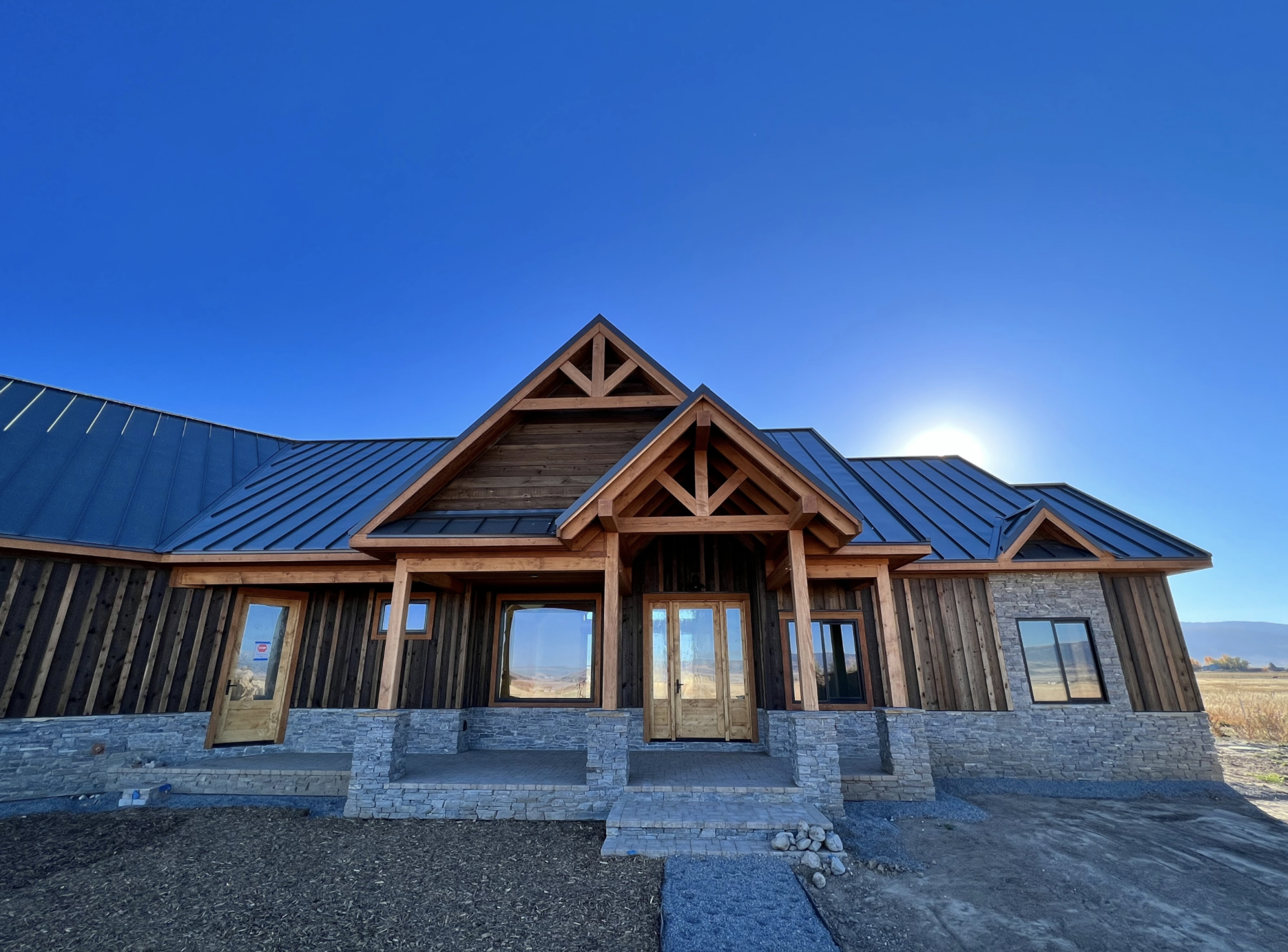 Newly constructed house with wooden and stone exterior, multiple gabled roofs, and large windows, under a clear blue sky.