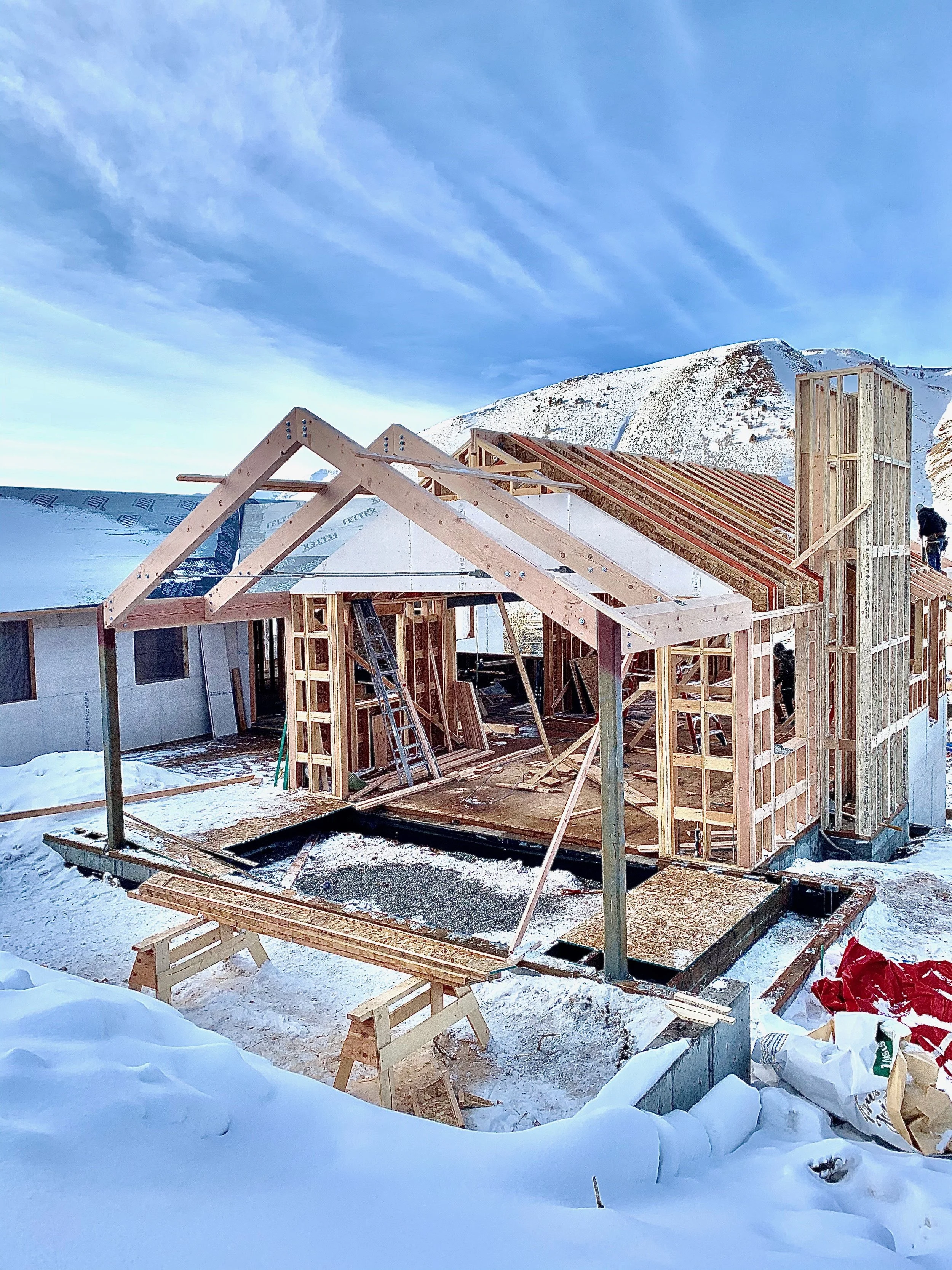 Construction site of a house in snowy landscape with a mountain in the background. Wooden framework and partially built roof are visible, with workers on site.