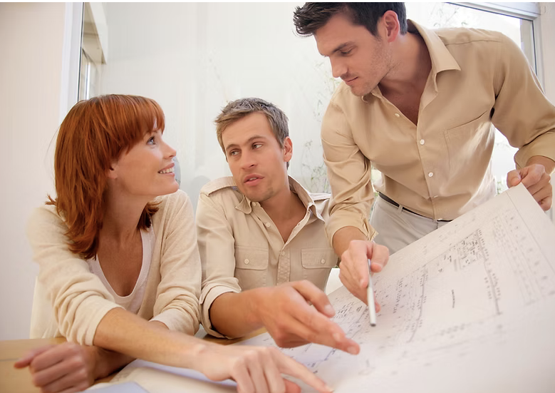 Three people working together on architectural blueprints, with one person pointing at the plans, in an office setting.