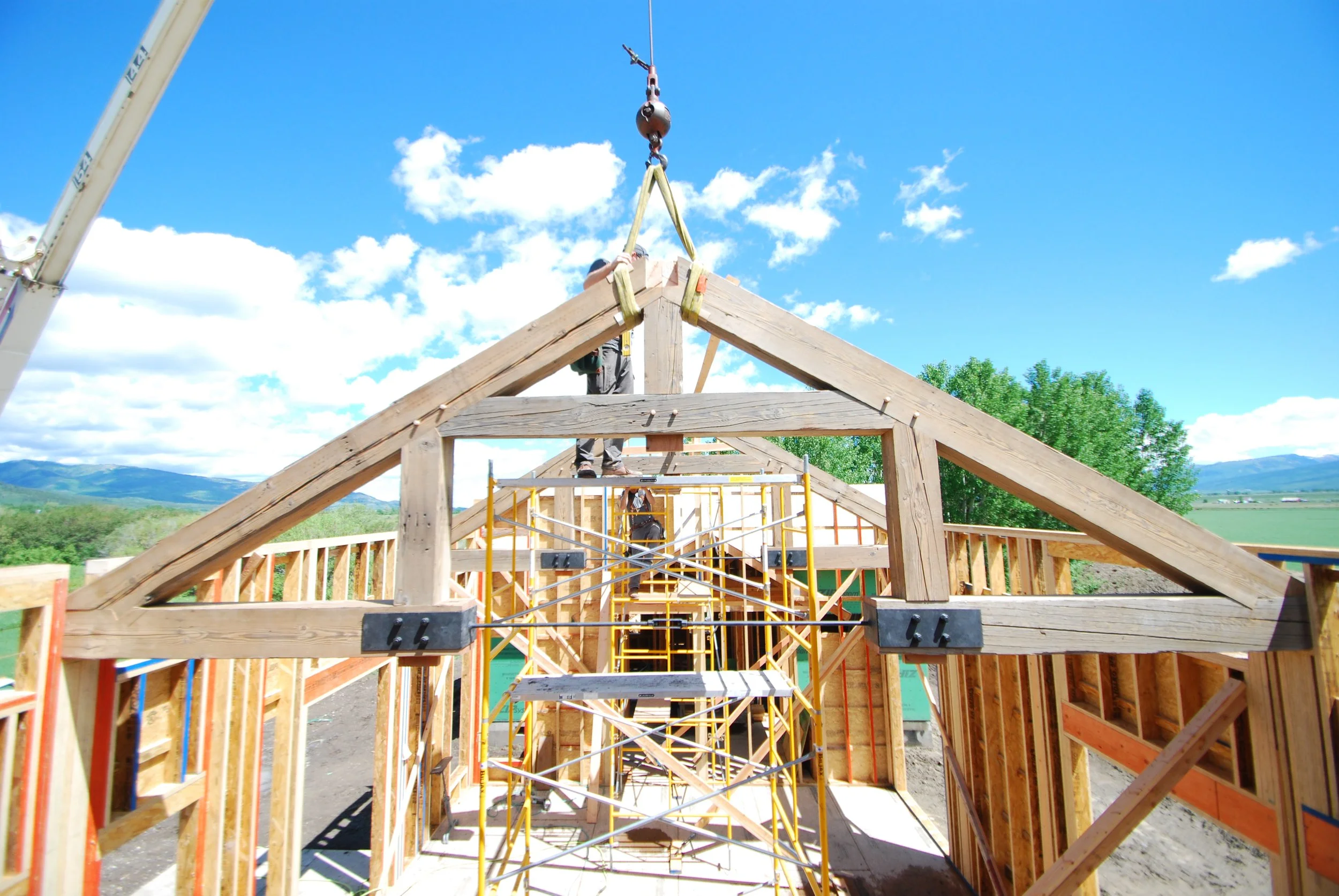 Construction of a wooden house frame outdoors under a blue sky with clouds, with scaffolding and workers visible.
