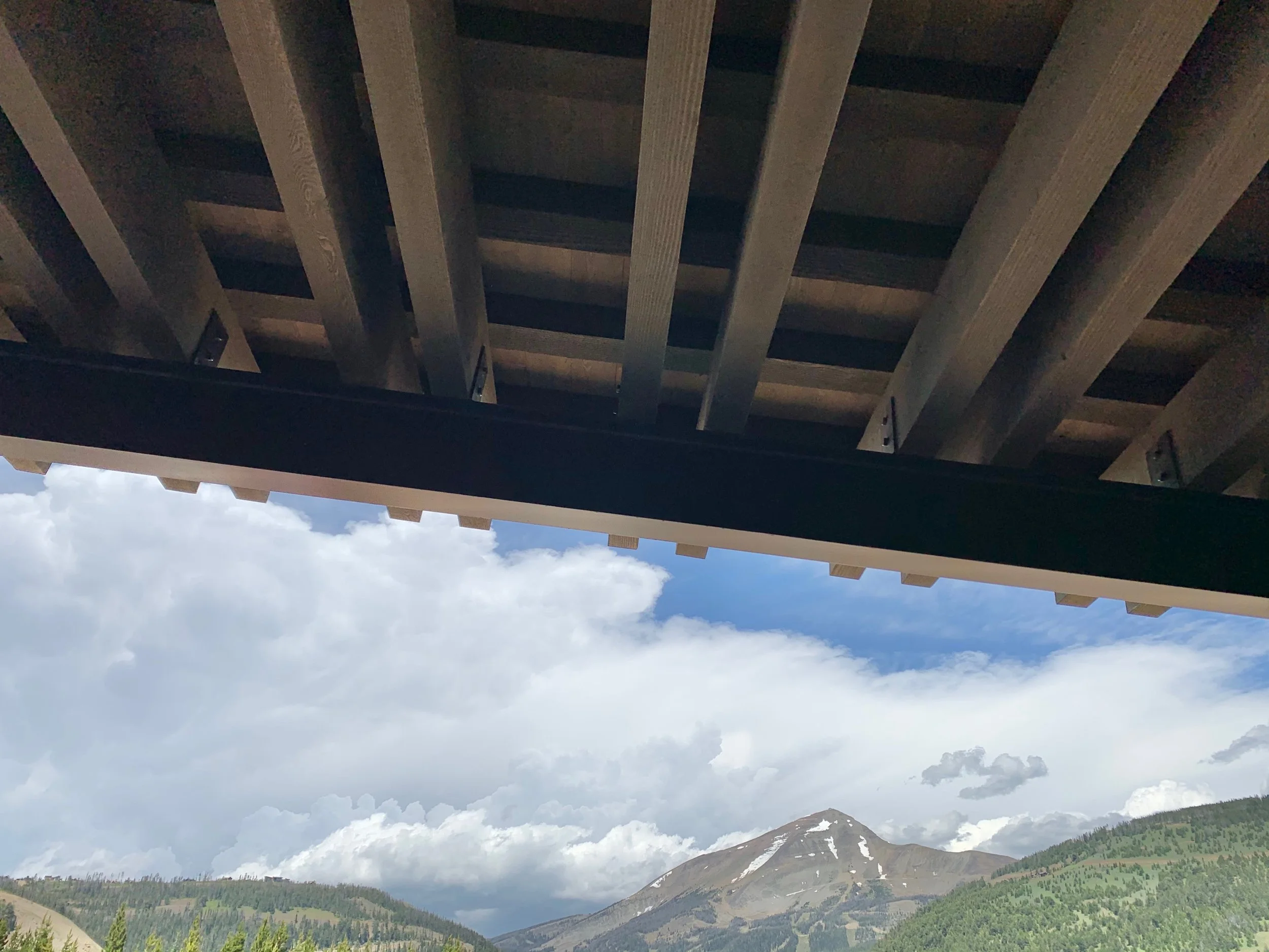 Looking up from beneath a wooden bridge or deck with a mountain and partly cloudy sky in the background.