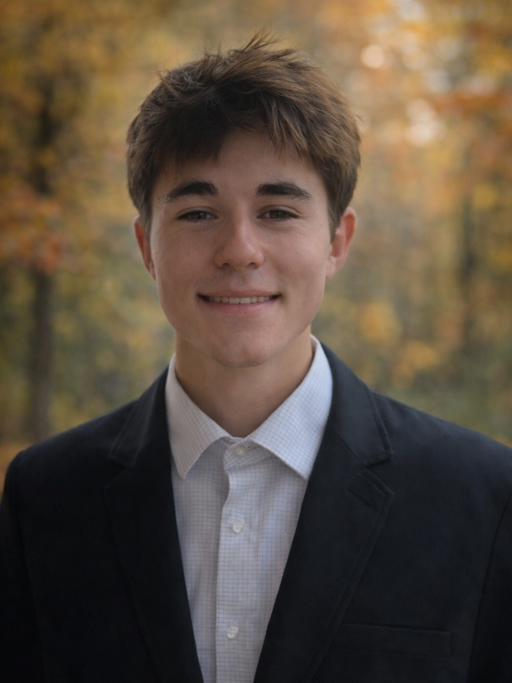 Young man in a black blazer and white shirt smiling outdoors with autumn trees in the background.