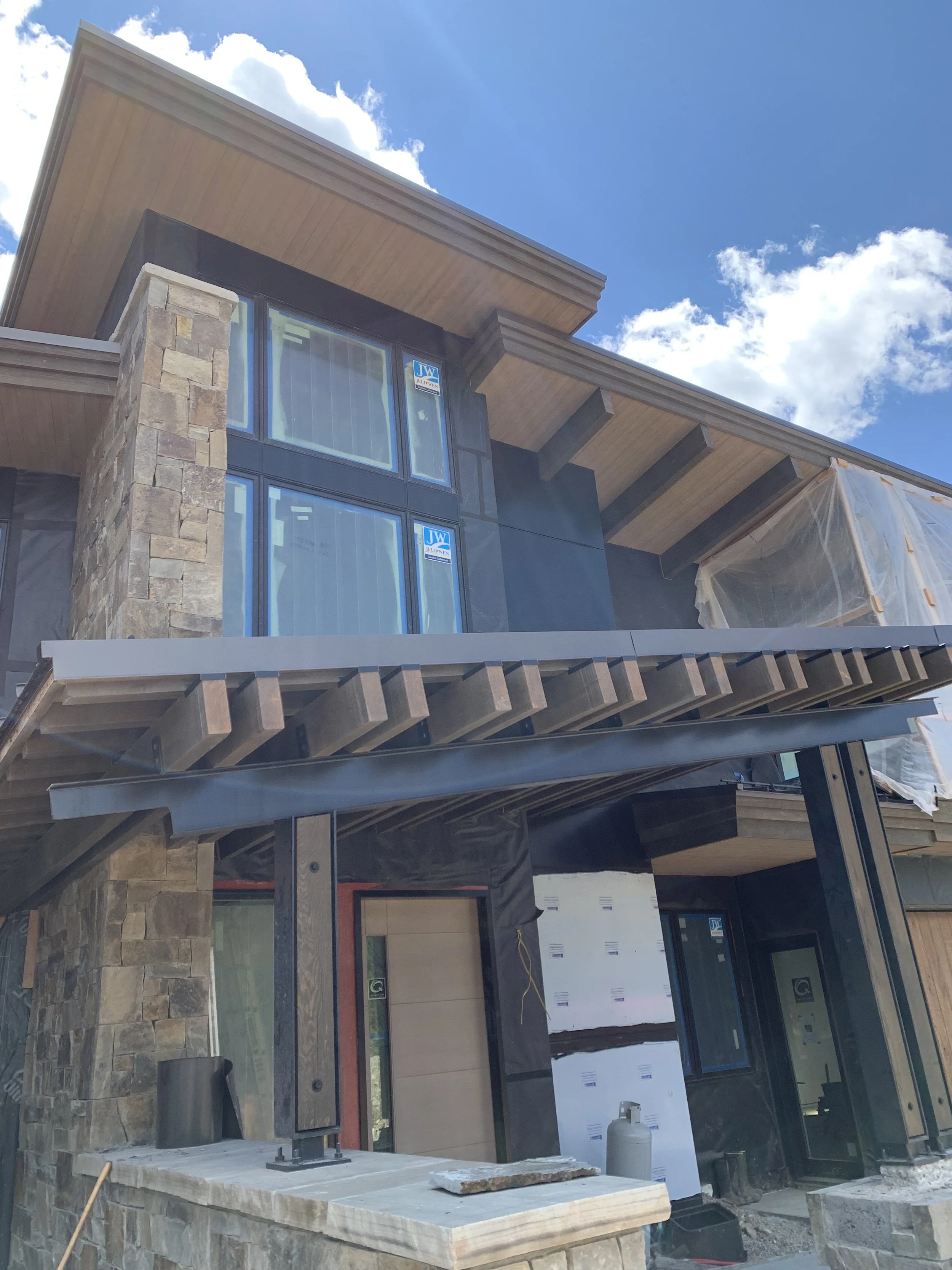 A modern multi-story house under construction with stone and wood exterior, large windows, and a partially installed deck, set against a blue sky with scattered clouds.