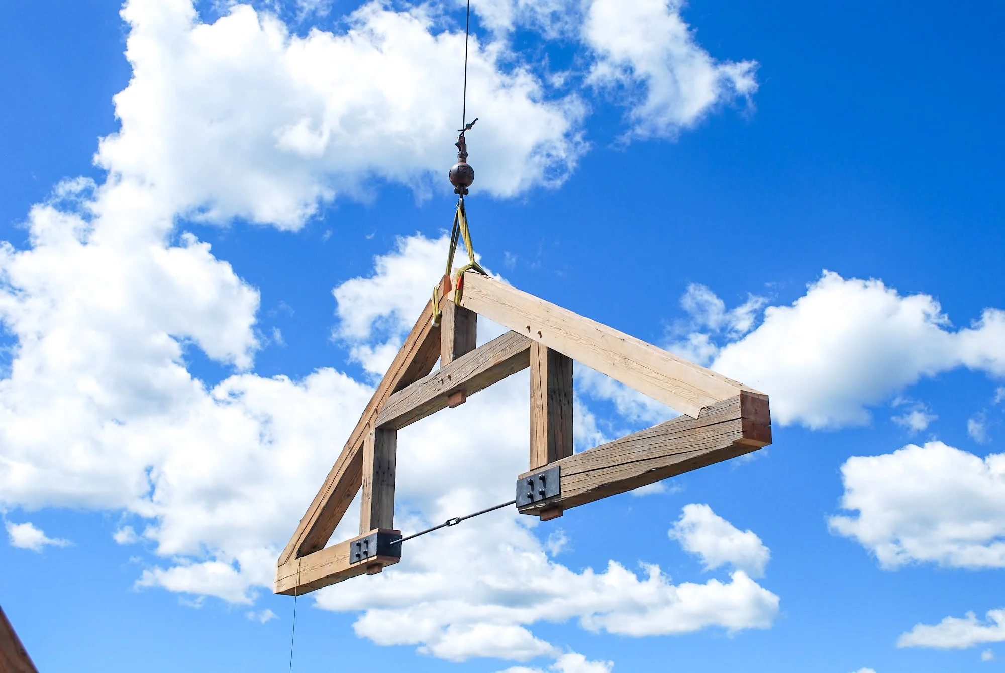 A wooden frame structure being lifted by a crane against a blue sky with white clouds.