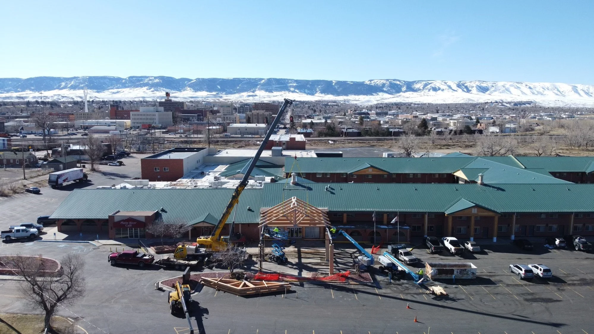 Construction workers installing a wooden roof structure on a building with green metal roof, construction vehicles and cranes, in a commercial area with mountains in the background.