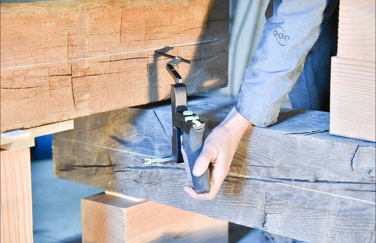 A person working with a pneumatic nail gun in a woodworking shop, installing a large wooden beam.