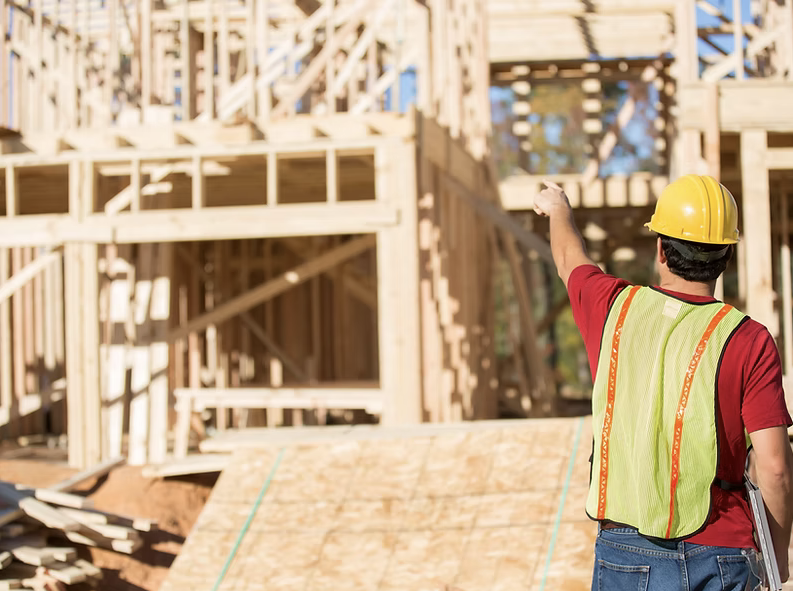 A construction worker wearing a yellow hard hat and a safety vest pointing at a wooden building under construction.
