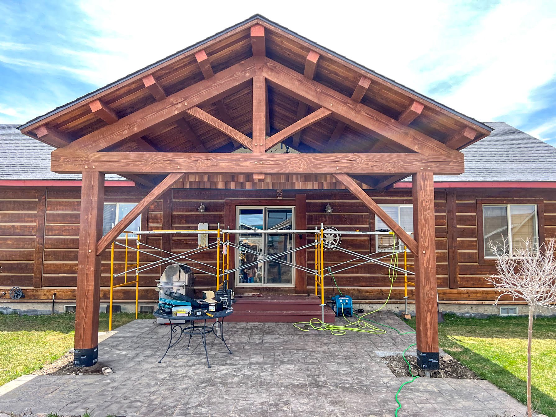 Backyard patio under construction with a new wooden porch and scaffolding, tools, and a small table with equipment in front.