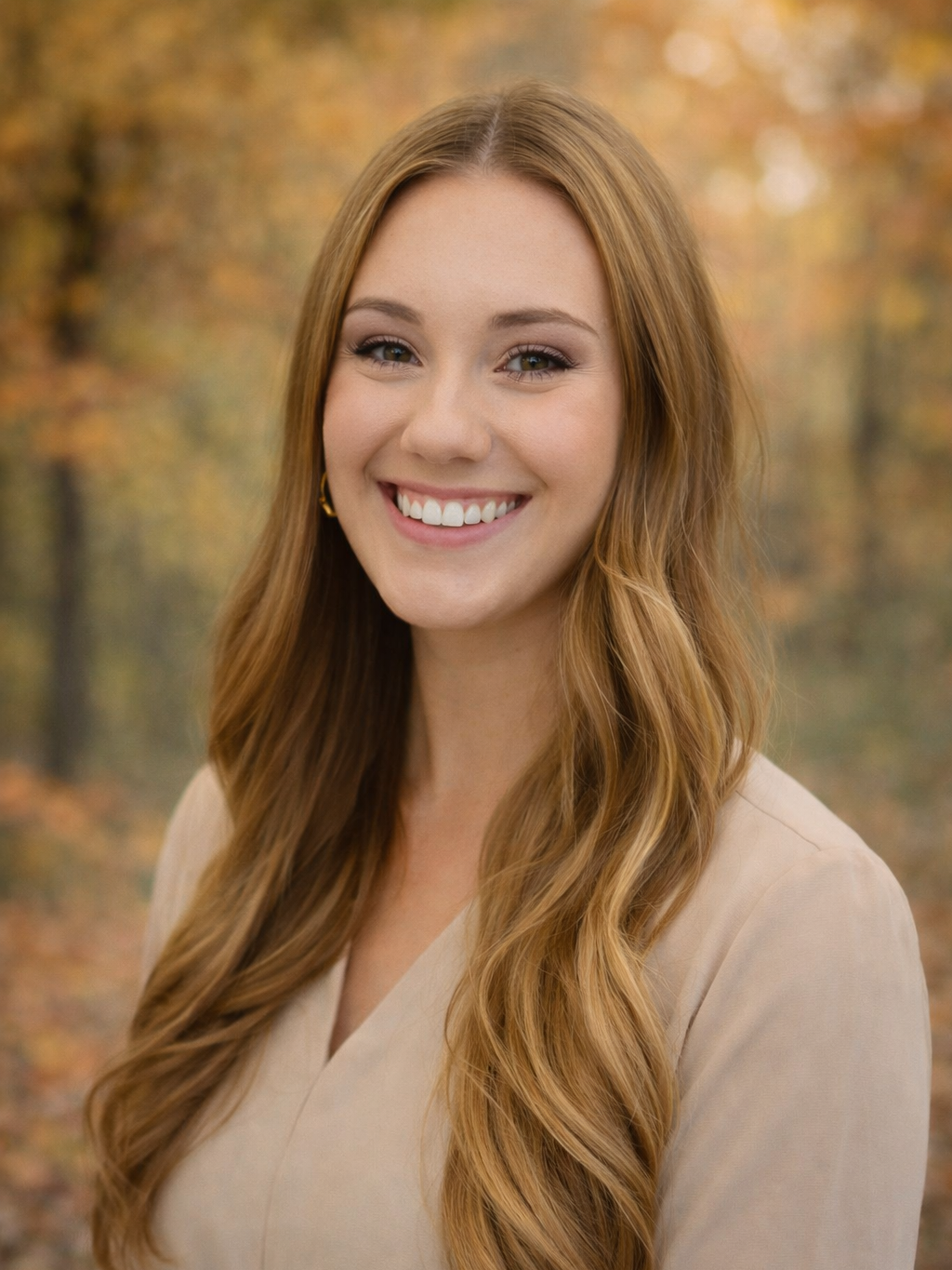 Young woman with long wavy red hair smiling outdoors in fall, wearing a light beige top and gold hoop earrings.